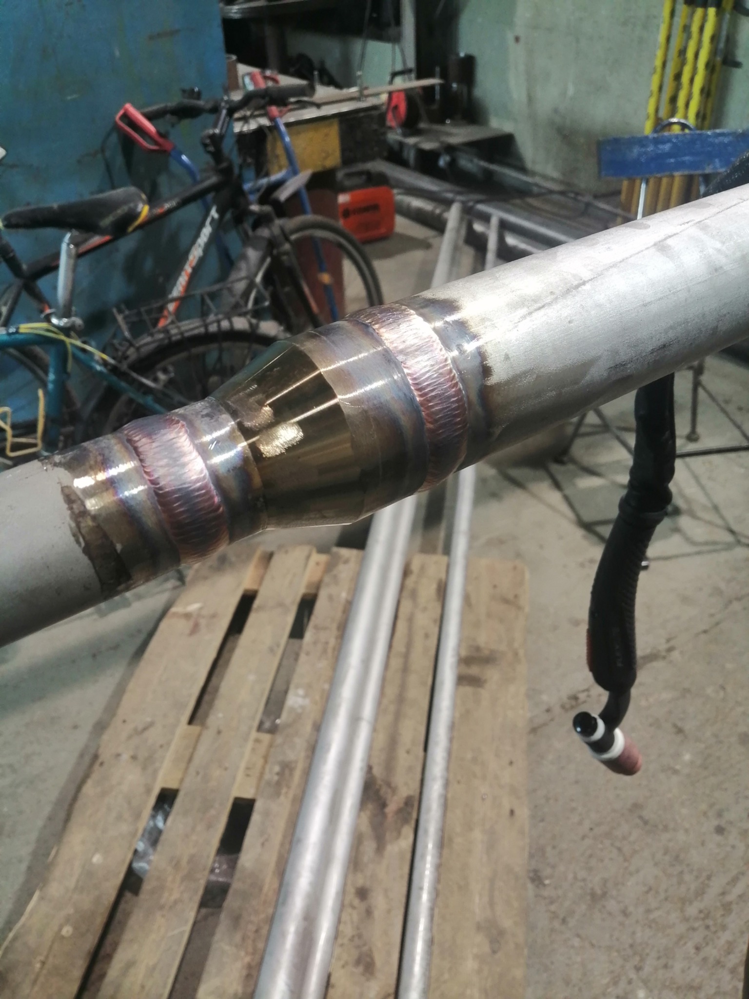 Spoina na rurze ze stali nierdzewnej, widoczne tęczowe przebarwienia po spawaniu TIG, w tle warsztat z rowerem i narzędziami.