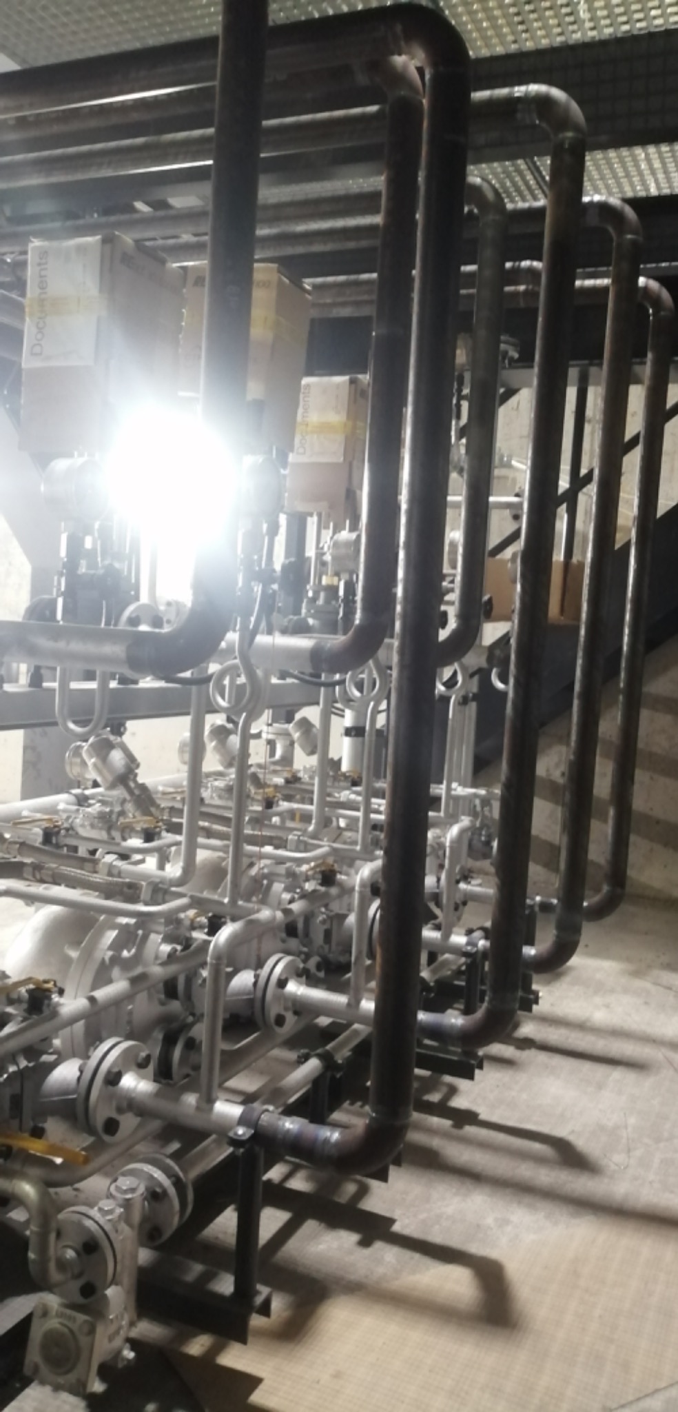 Instalacja przemysłowa z rurami i zaworami, widoczne spawy na rurach, oświetlenie górne powoduje silne odblaski.