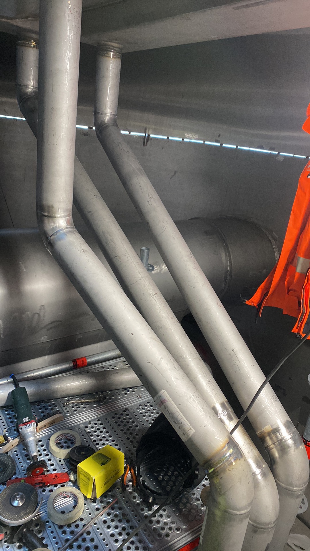 Wnętrze metalowej konstrukcji z trzema rurami ze stali nierdzewnej połączonymi spawami, widoczne narzędzia szlifierskie i ochronna odzież robocza.