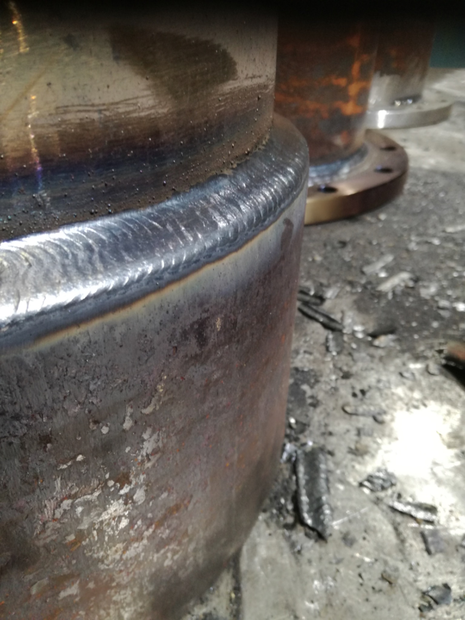 Szczegółowe zbliżenie spoiny na metalowej rurze z widocznymi śladami obróbki i odpryskami na podłożu.