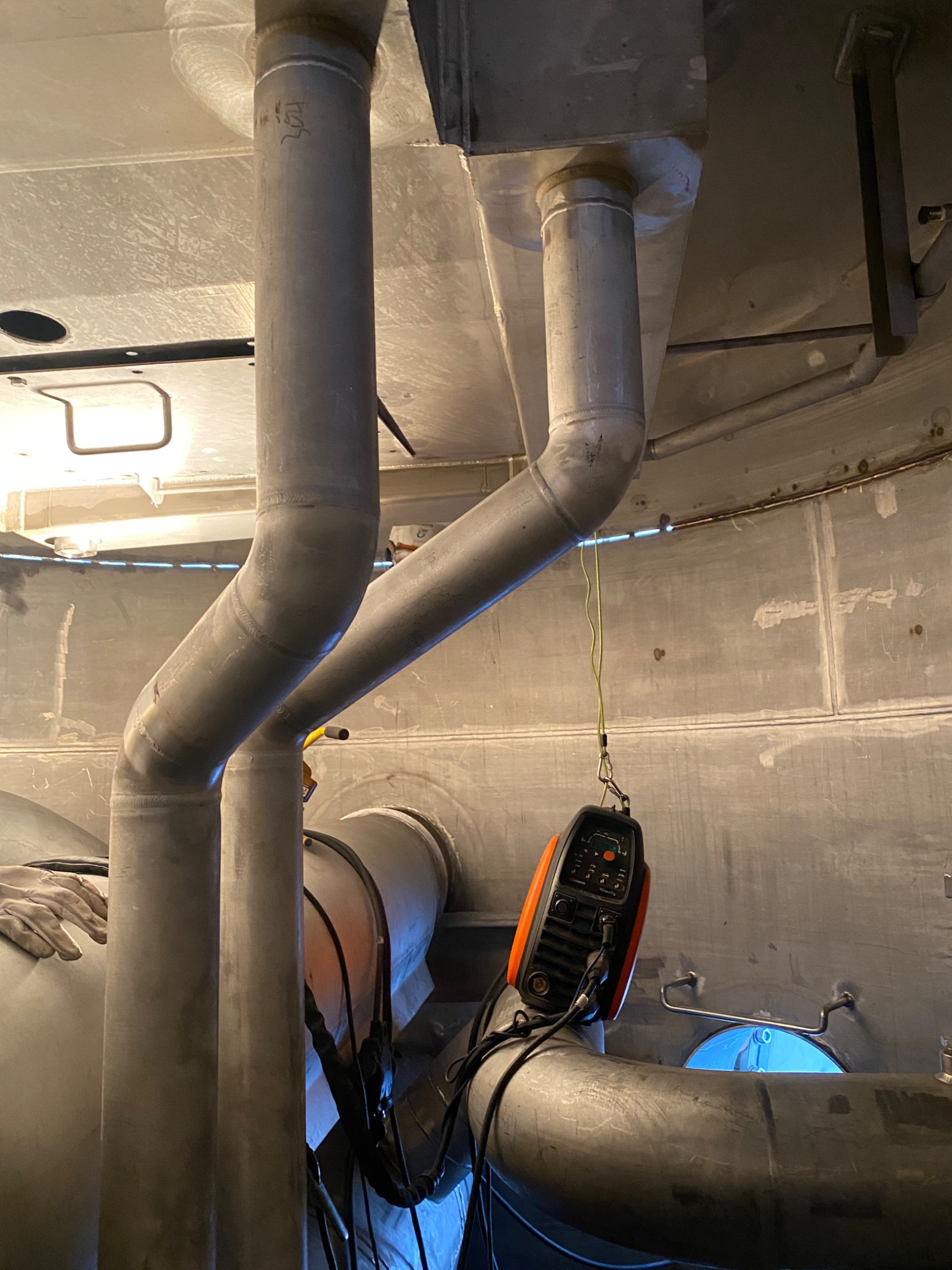 Wnętrze konstrukcji metalowej z grubymi rurami, widoczny migający łuk elektryczny podczas spawania rur, wisząca spawarka inwertorowa z pomarańczowymi elementami obudowy.