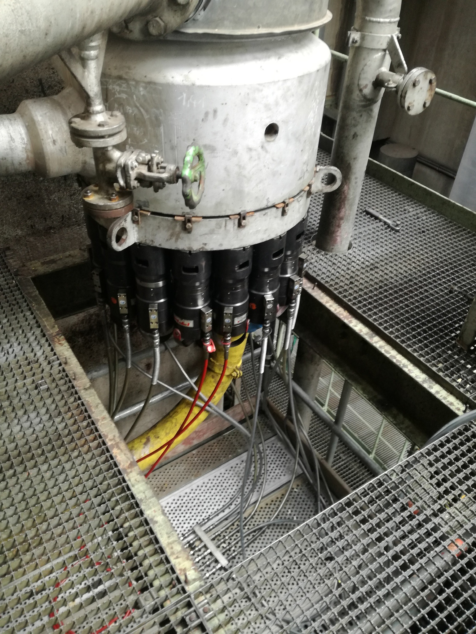 Metalowa konstrukcja przemysłowa z rurami, zaworami z zielonym pokrętłem i cylindrycznymi elementami z przewodami, umieszczona na metalowej platformie kratowej.