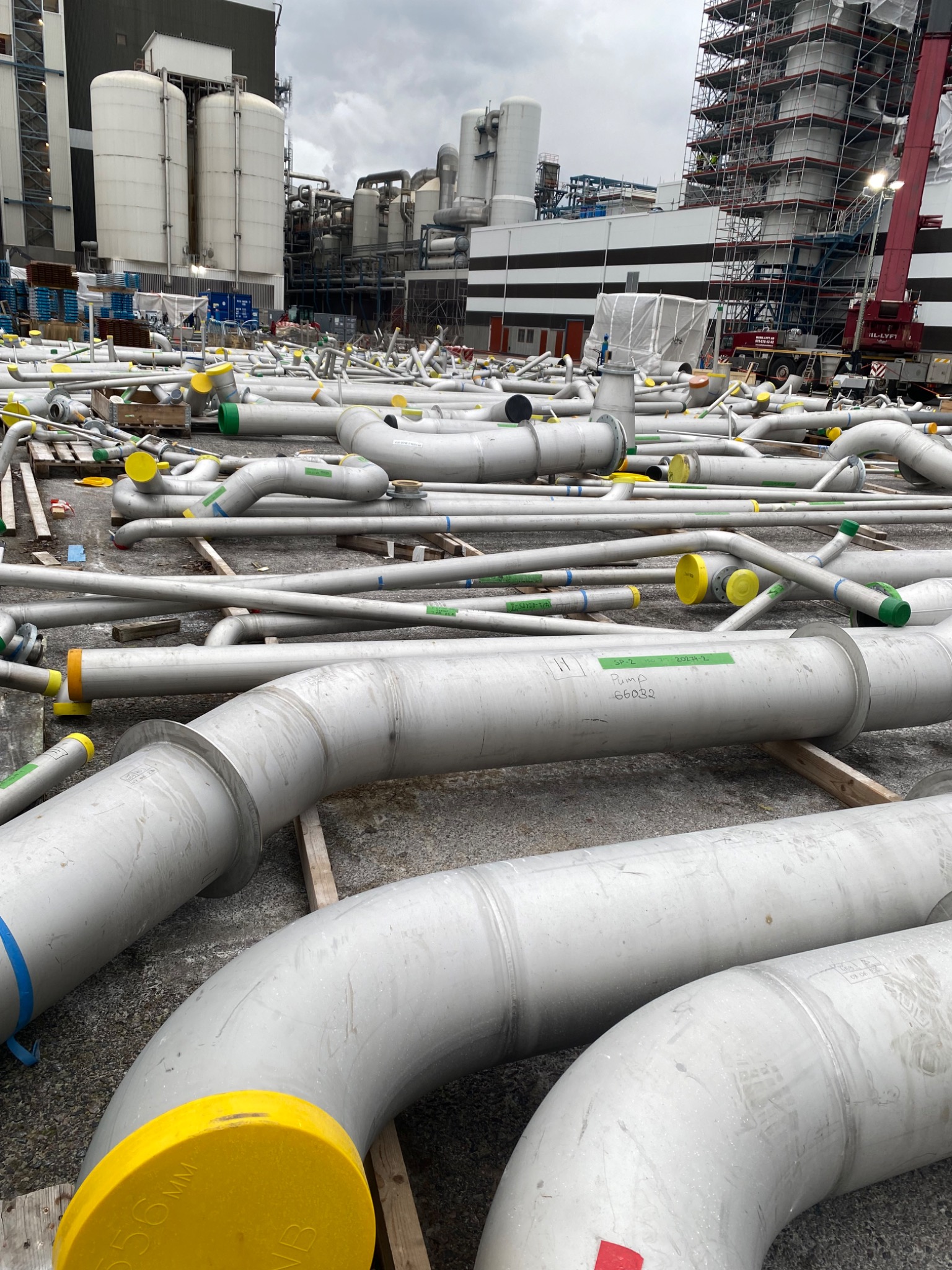 Srebrne rury przemysłowe z kolorowymi zaślepkami, ułożone na placu budowy z widocznymi zbiornikami i rusztowaniami w tle, oczekujące na montaż.