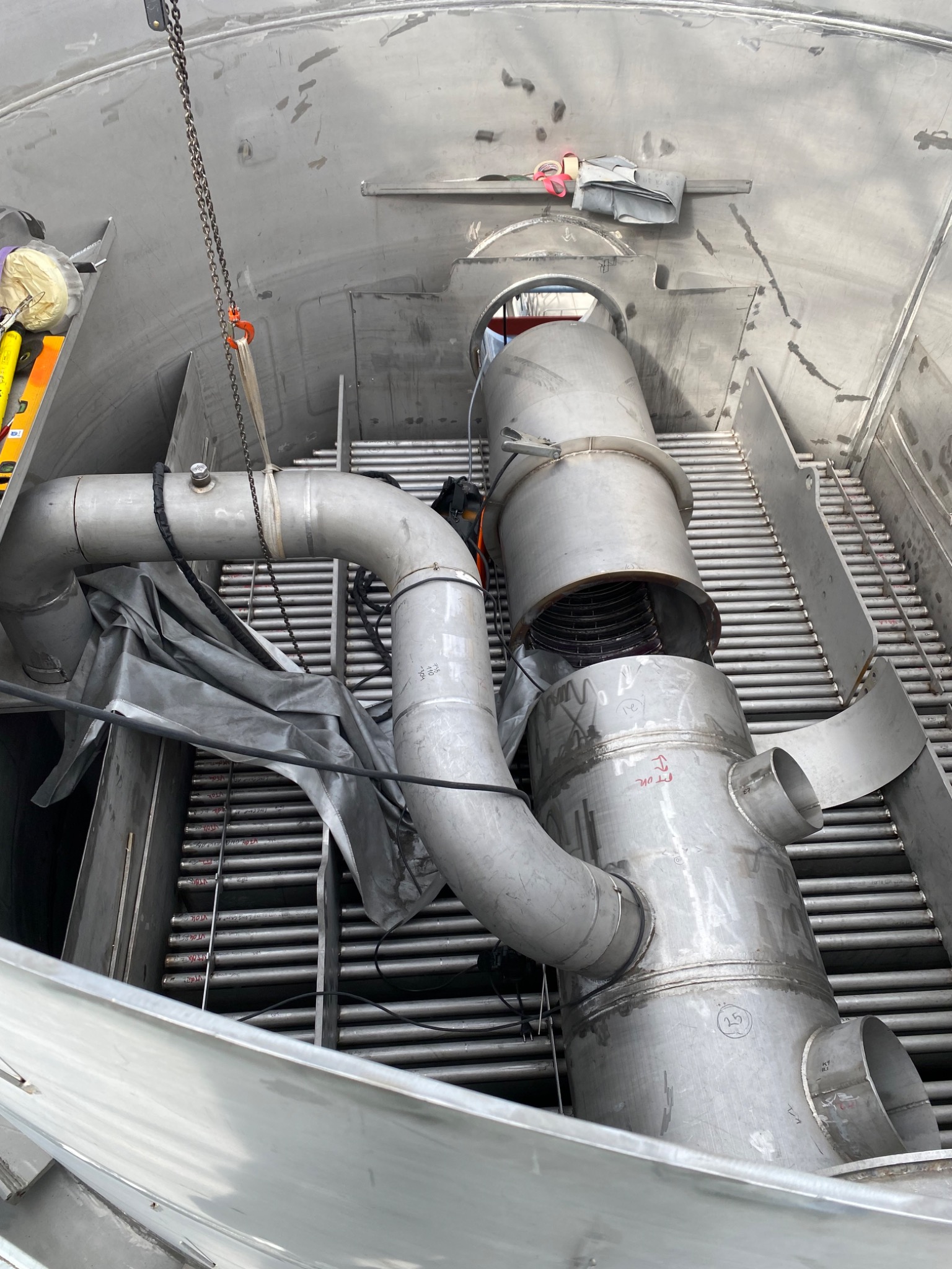 Wnętrze metalowej konstrukcji z rurami i elementami podwieszonymi na łańcuchu, widoczne ślady obróbki i montażu.