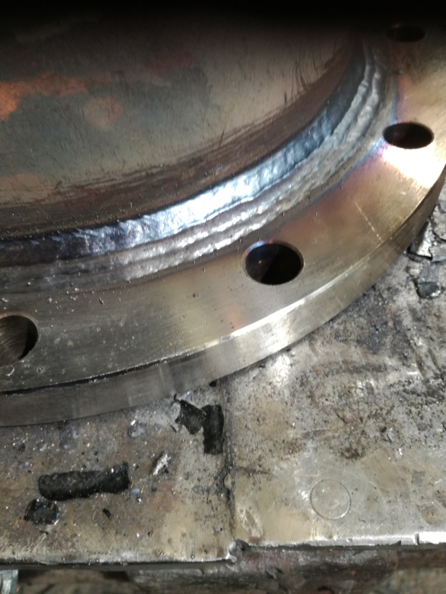 Spoina na metalowym kołnierzu z widocznymi otworami, leżącym na brudnym stole spawalniczym.