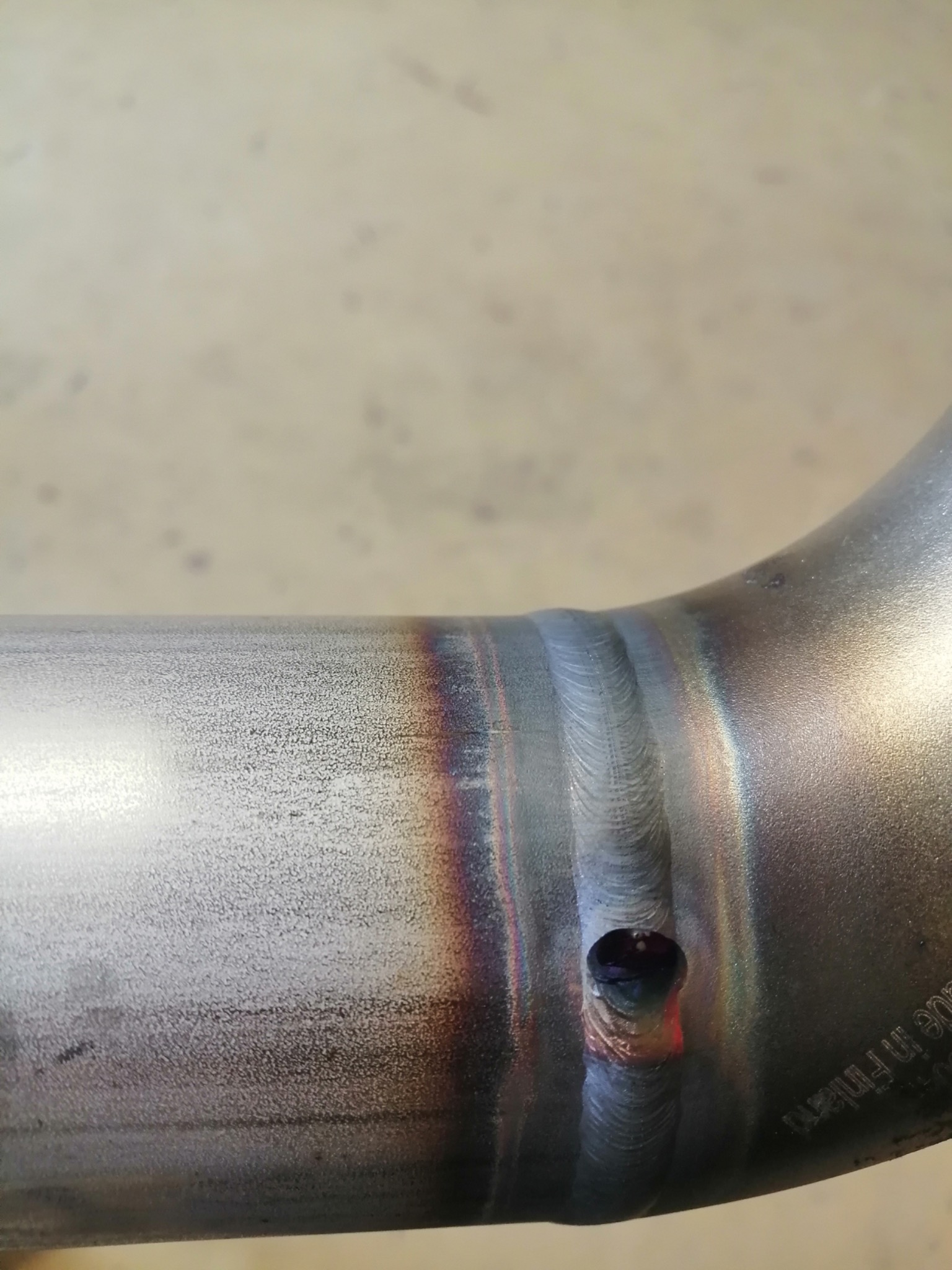 Detale spoiny na rurze ze stali nierdzewnej z otworem, widoczne przebarwienia termiczne i napis Made in Finland.