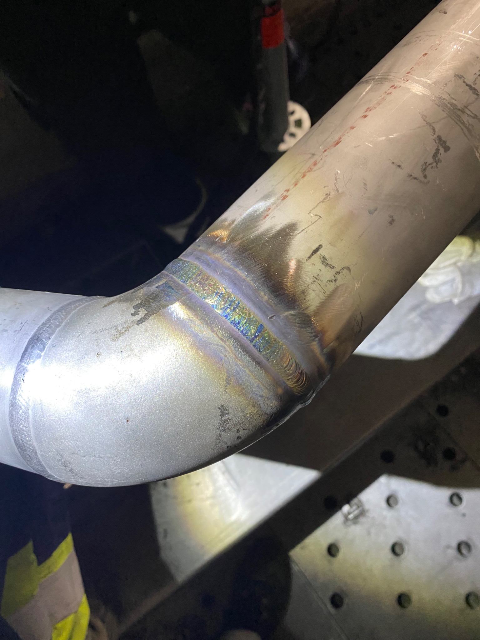 Spoina na rurze ze stali nierdzewnej, widoczne tęczowe przebarwienia po spawaniu, rura ułożona na metalowym stole spawalniczym.