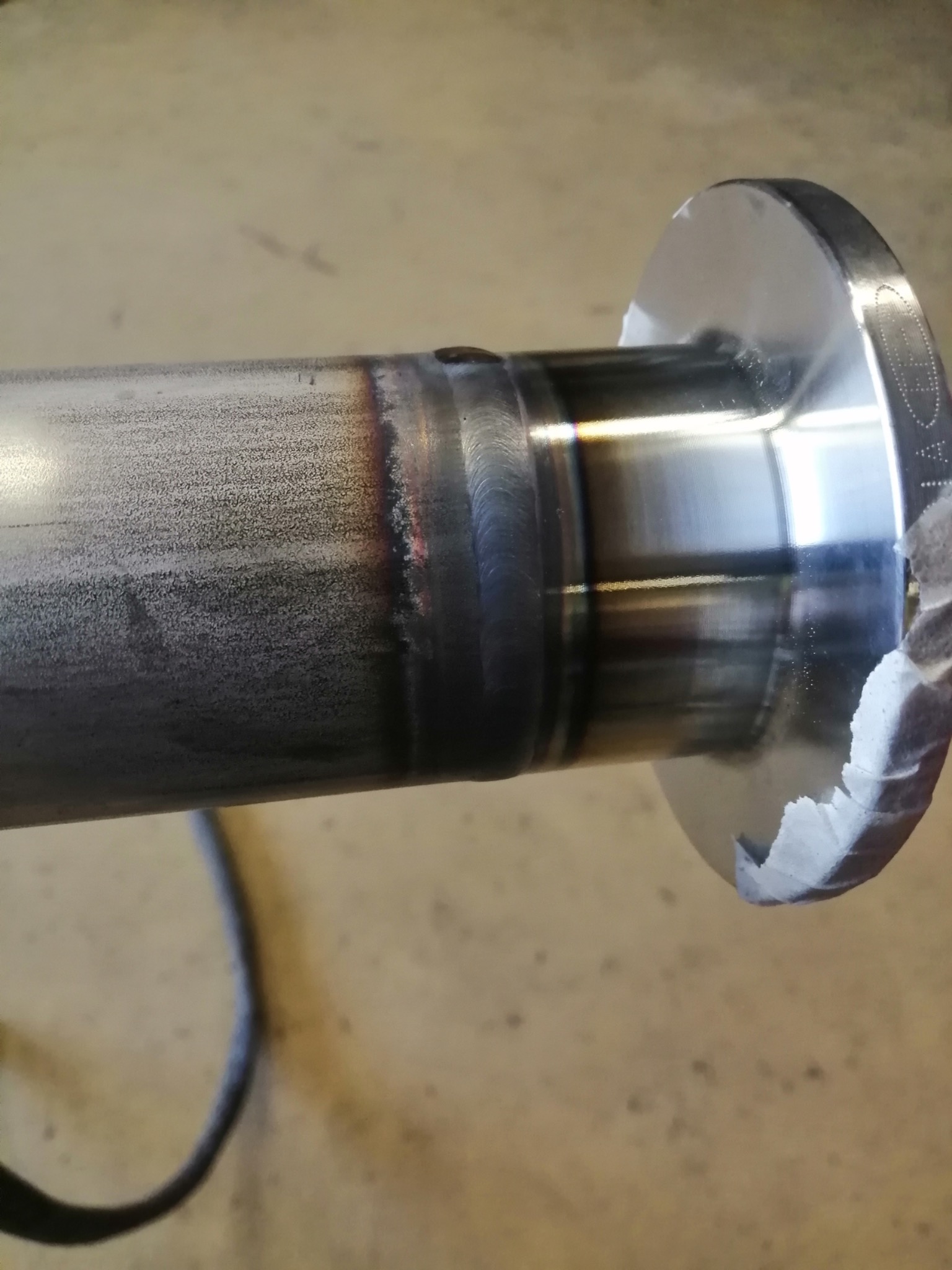 Spoina łącząca rurę z metalowym kołnierzem, widoczne ślady obróbki termicznej i niedoskonałości spoiny, fragment taśmy maskującej na kołnierzu.