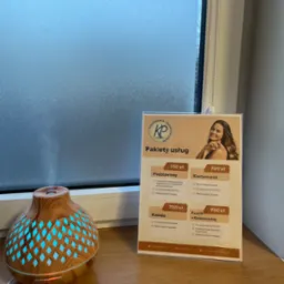 Aromaterapeutyczny dyfuzor obok oferty pakietów usług fizjoterapeutycznych z logo firmy, na parapecie okiennym z matową szybą.