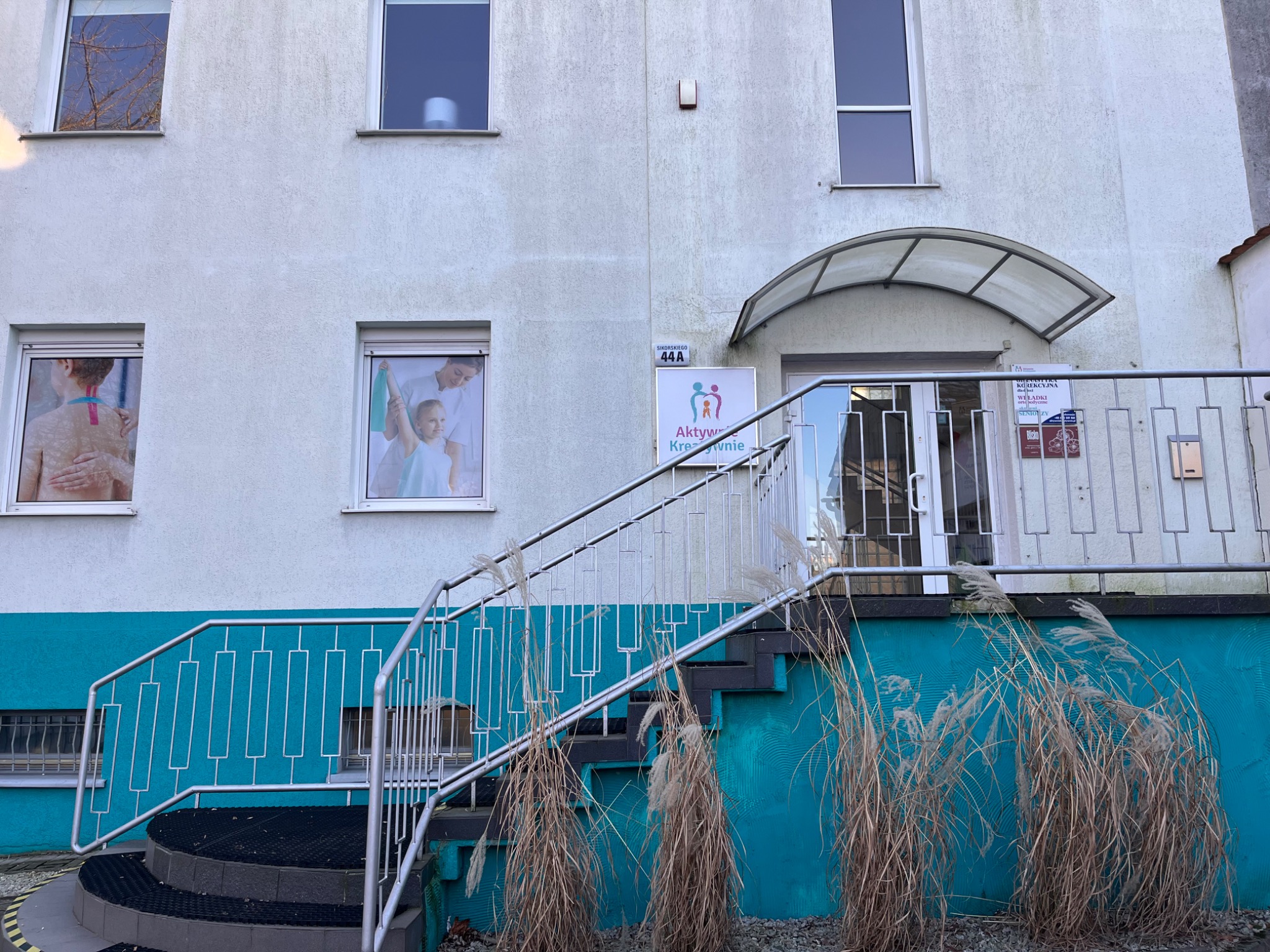 Fasada budynku z niebieskim cokołem, schodami z metalową barierką i szyldem 'Aktywnie Kreacyjnie'. W oknach widoczne plakaty przedstawiające zabiegi terapeutyczne.