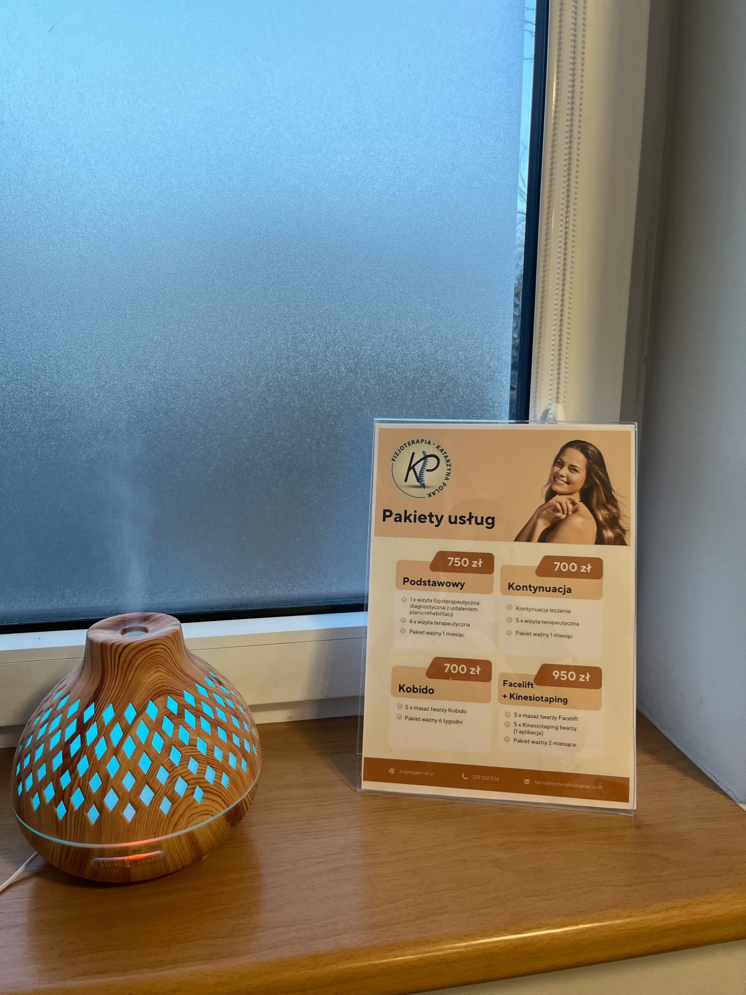 Aromaterapeutyczny dyfuzor obok oferty pakietów usług fizjoterapeutycznych z logo firmy, na parapecie okiennym z matową szybą.