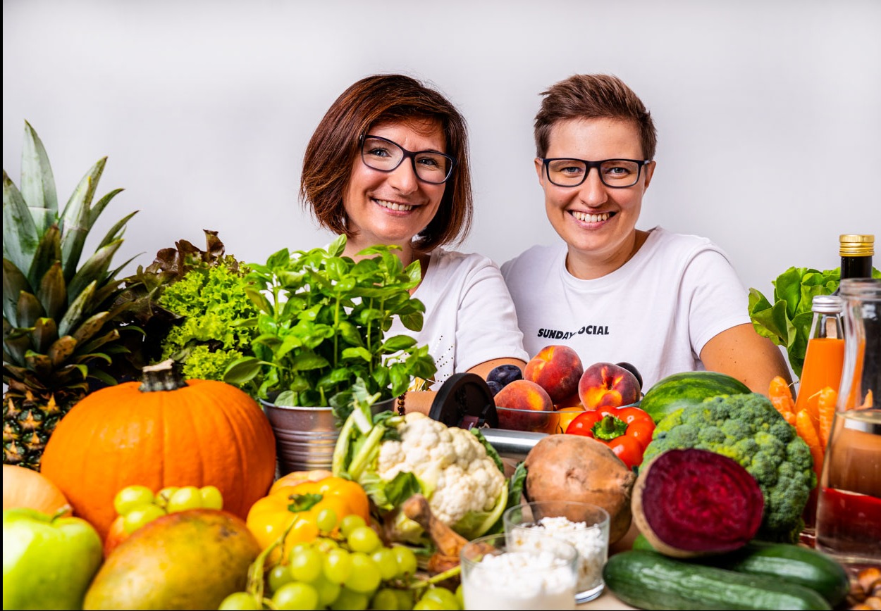 Dwie uśmiechnięte kobiety w okularach, otoczone różnorodnymi świeżymi owocami i warzywami, w tym ananasem, dynią, brokułami i burakami, oraz domowymi sokami w szklanych butelkach.