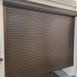 Zasunięta brązowa roleta zewnętrzna z widoczną czarną wycieraczką i fragmentem podłogi wewnątrz pomieszczenia.