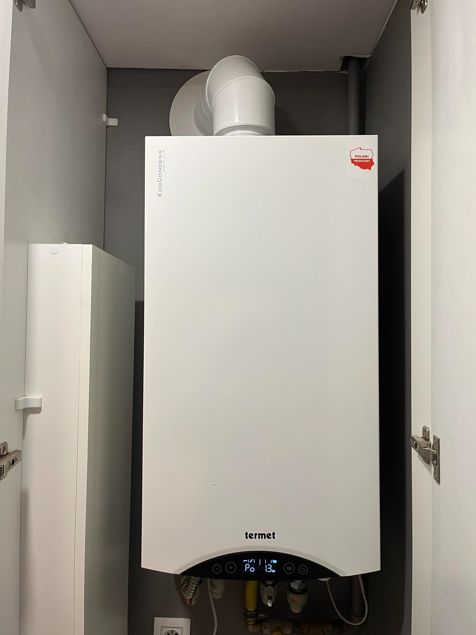 Biały piec gazowy Termet EcoCondens Gold Plus, zamontowany w szafce, widoczny ekran z parametrami pracy i naklejka 'Polski Producent'.
