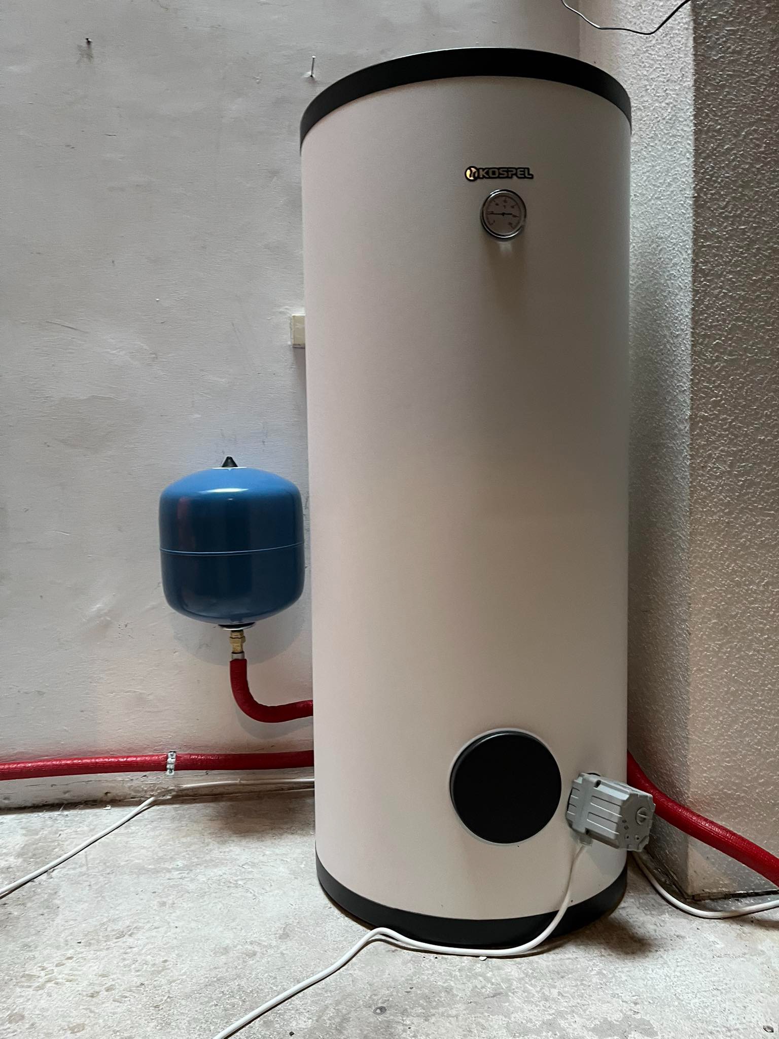 Biały, cylindryczny bojler marki Kospel z manometrem i podłączonym niebieskim zbiornikiem wyrównawczym, zamontowany na szarej ścianie z widocznymi czerwonymi rurami instalacji grzewczej.