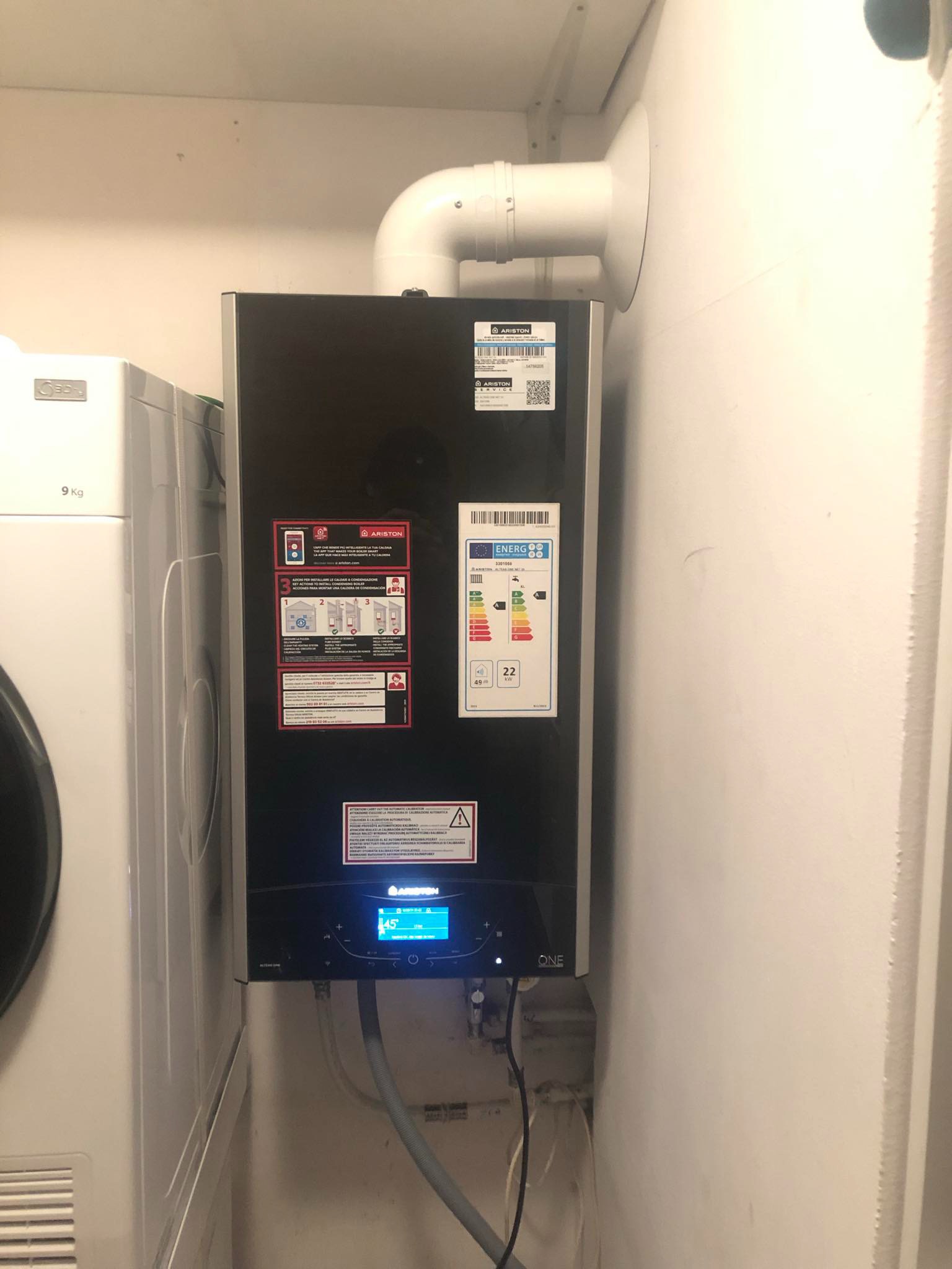 Zamontowany piec gazowy Ariston ONE z podłączonymi przewodami, obok pralka, widoczne etykiety i instrukcje na obudowie.