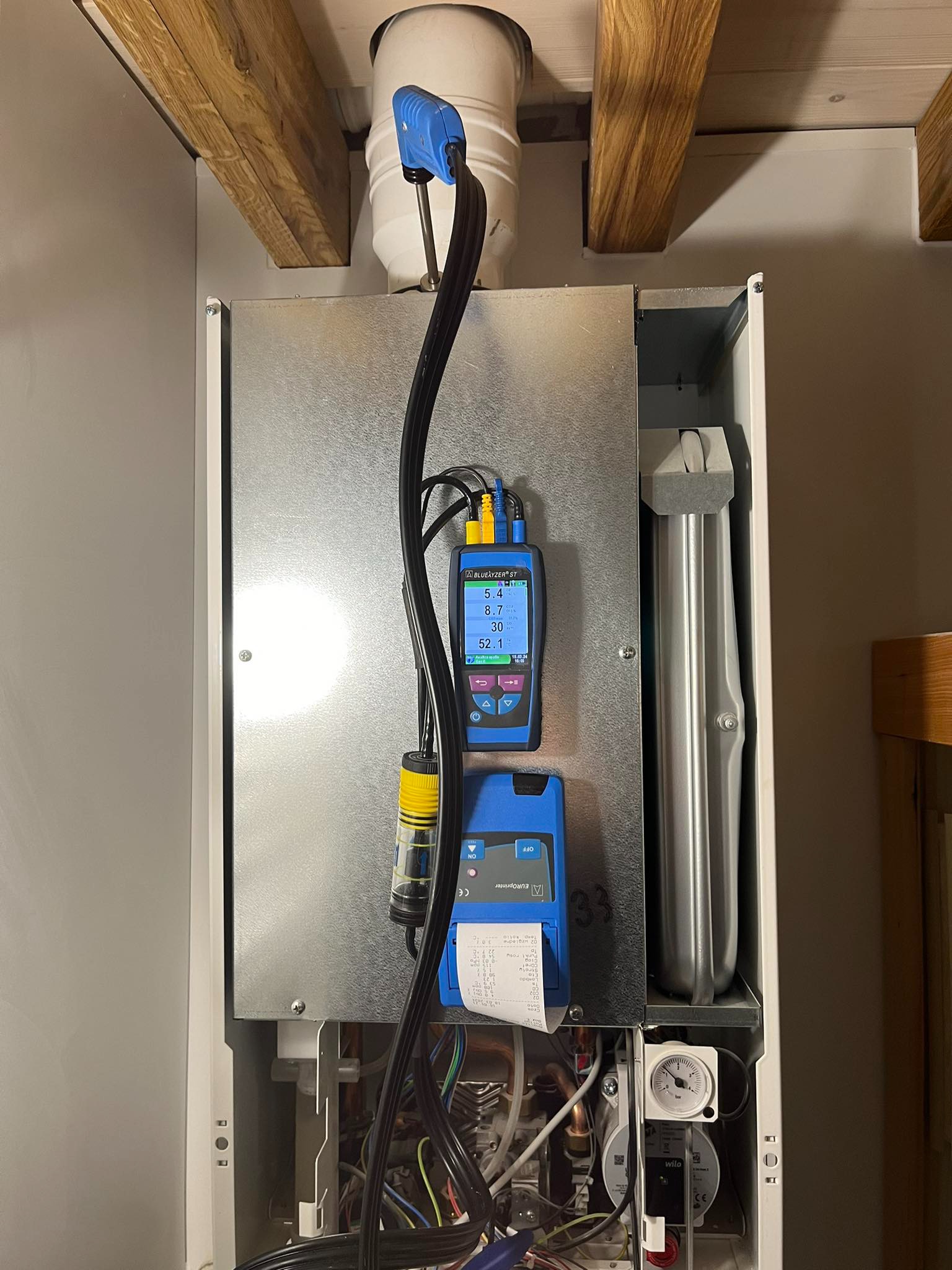 Analizator spalin podłączony do pieca gazowego, wyświetlający wyniki pomiarów na ekranie LCD. Widoczny wydruk z pomiarów oraz elementy instalacji grzewczej.