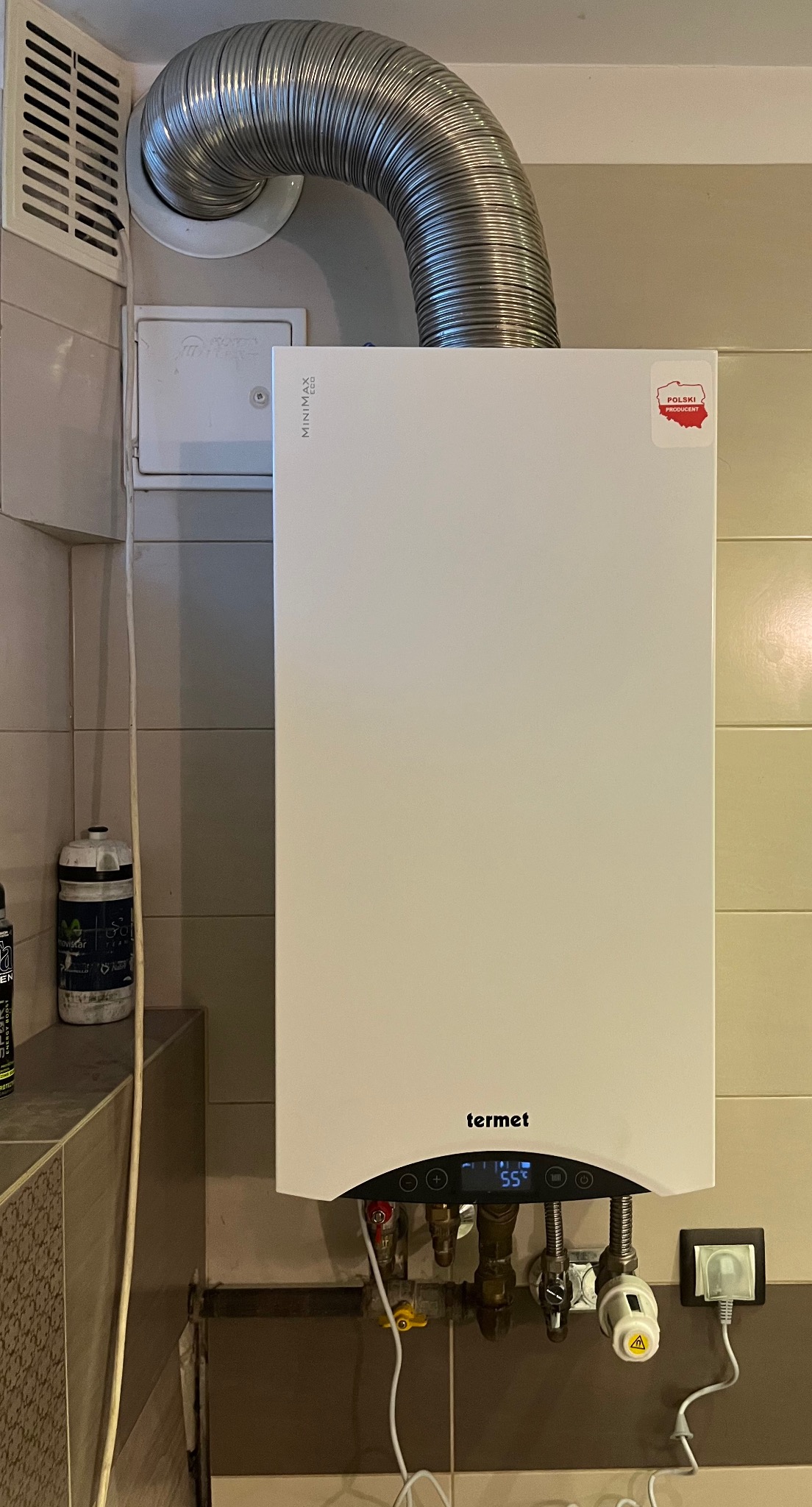Biały piec gazowy Termet z podłączonym przewodem odprowadzającym spaliny, zamontowany na ścianie wyłożonej płytkami, widoczny wskaźnik temperatury 55 stopni.