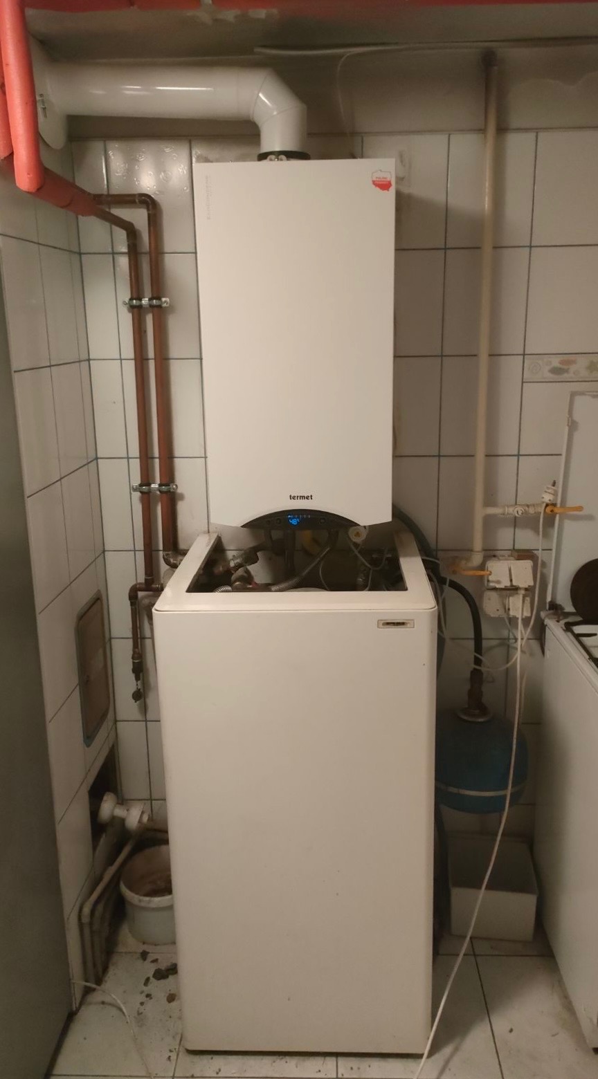 Biały piec gazowy Termet z otwartą obudową w trakcie montażu, widoczne rury miedziane i białe płytki ceramiczne na ścianach.