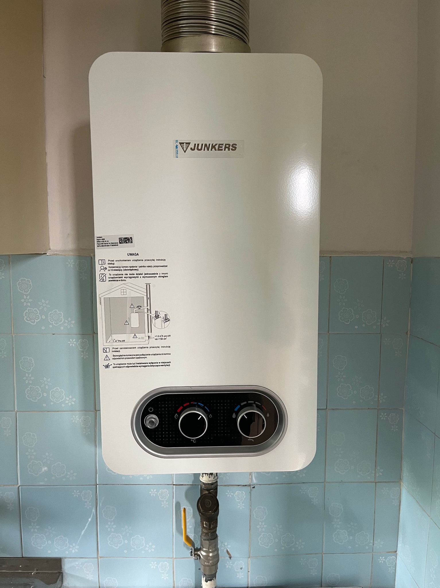 Biały piec gazowy Junkers zamontowany na ścianie z niebieskich płytek z kwiatowym wzorem, widoczne przyłącze gazowe z żółtym zaworem, instrukcje bezpieczeństwa na obudowie pieca.