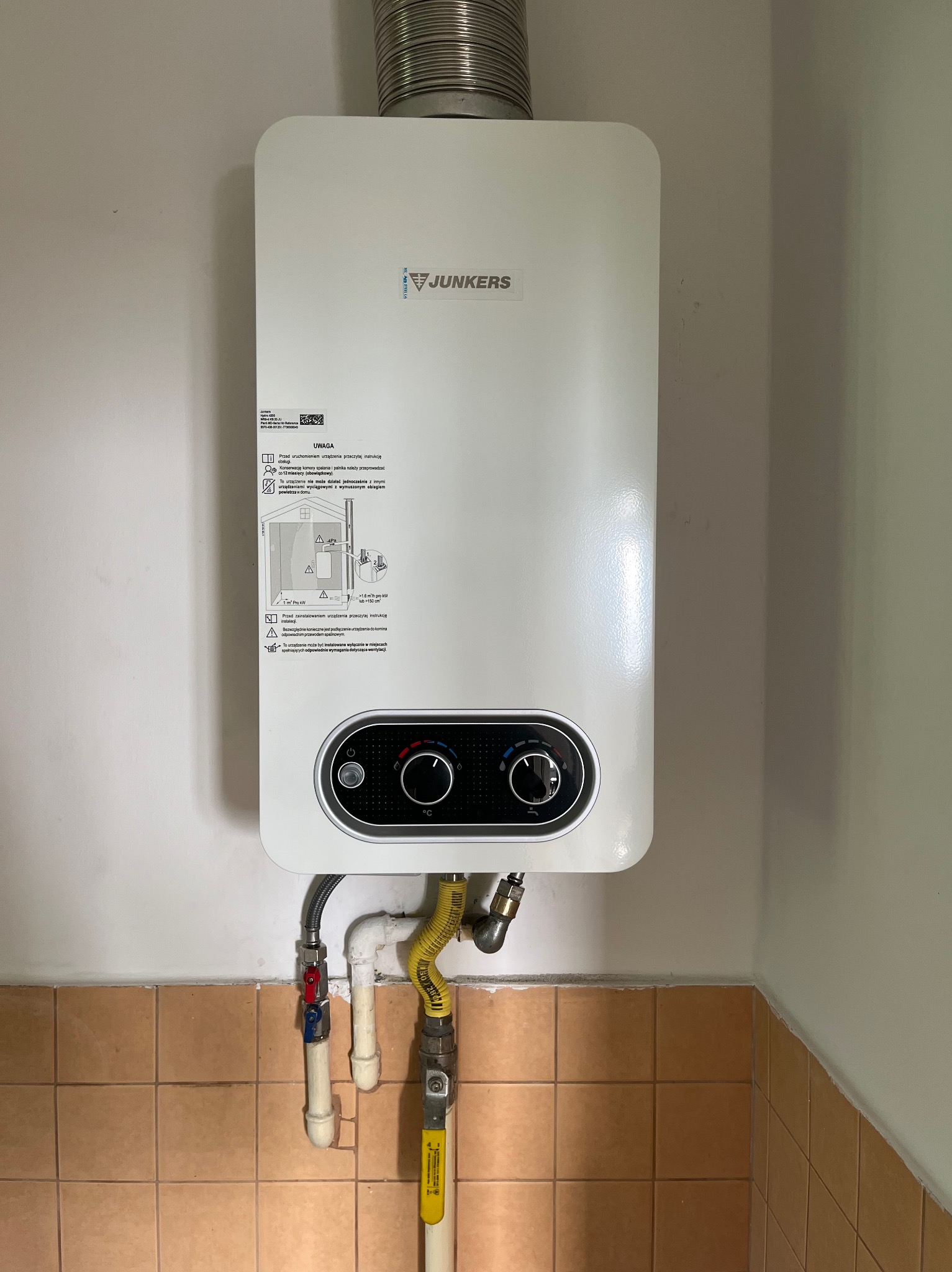 Nowy, biały piec gazowy Junkers zamontowany na ścianie w Krakowie, z widocznymi przyłączami gazowymi i wodnymi oraz rurą odprowadzającą spaliny.