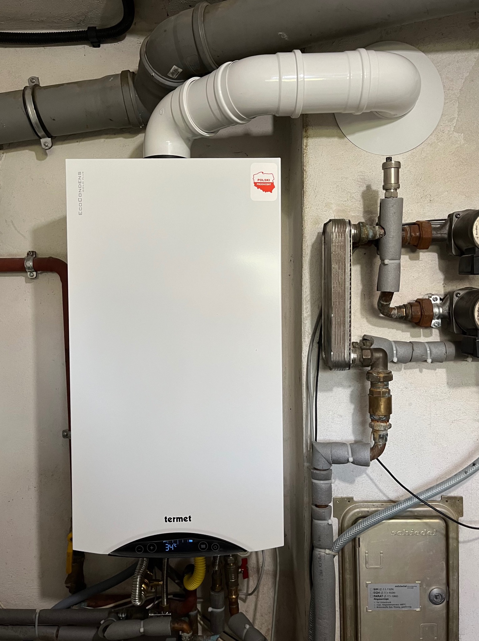 Biały kondensacyjny piec gazowy Termet z widocznymi rurami odprowadzającymi spaliny, zamontowany na ścianie z widocznymi elementami instalacji grzewczej i oznaczeniem 'Polski Producent'.