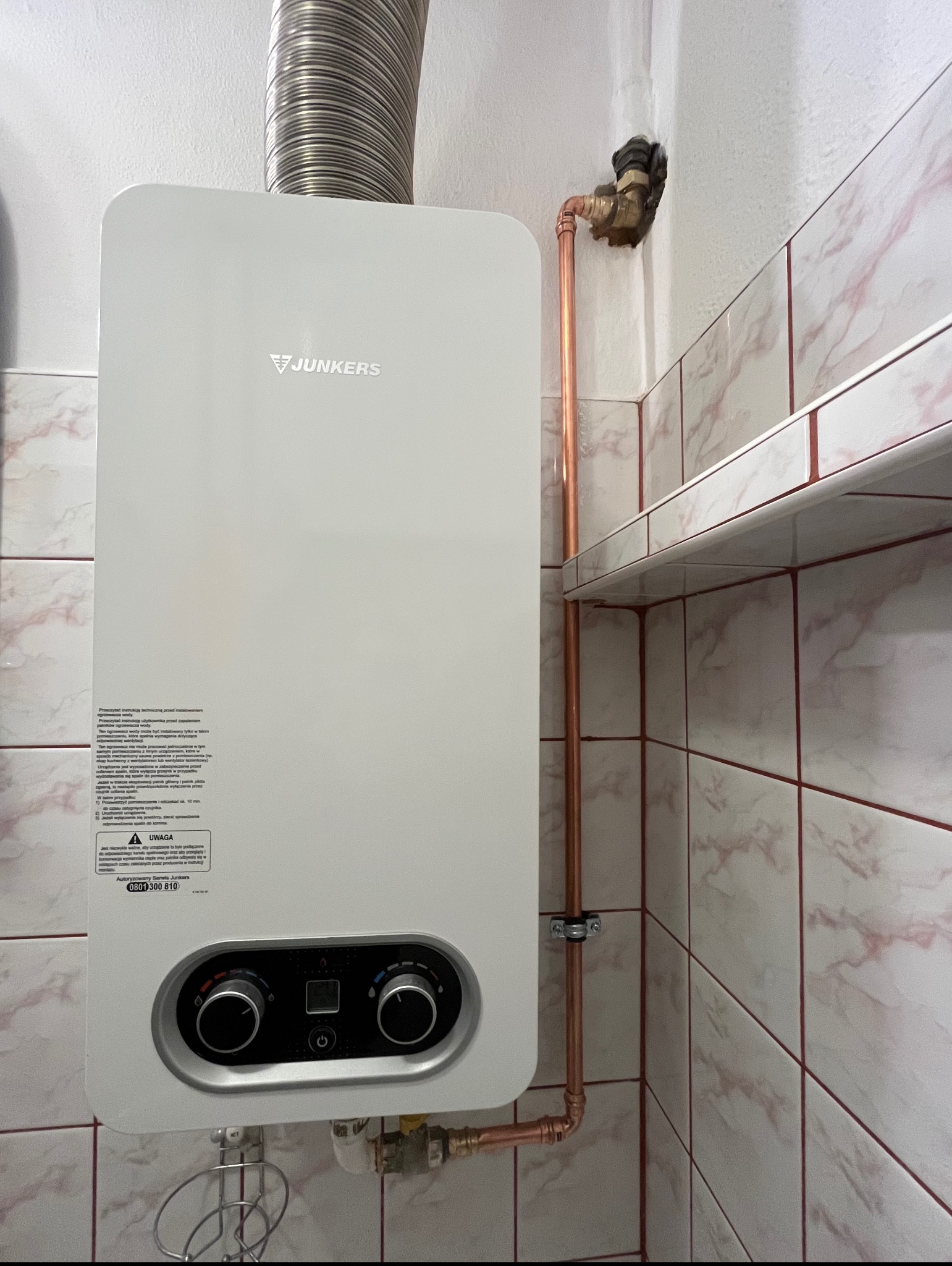 Biały piec gazowy Junkers zamontowany na ścianie z marmurowymi płytkami, widoczne miedziane rury instalacji gazowej i rura odprowadzająca spaliny.