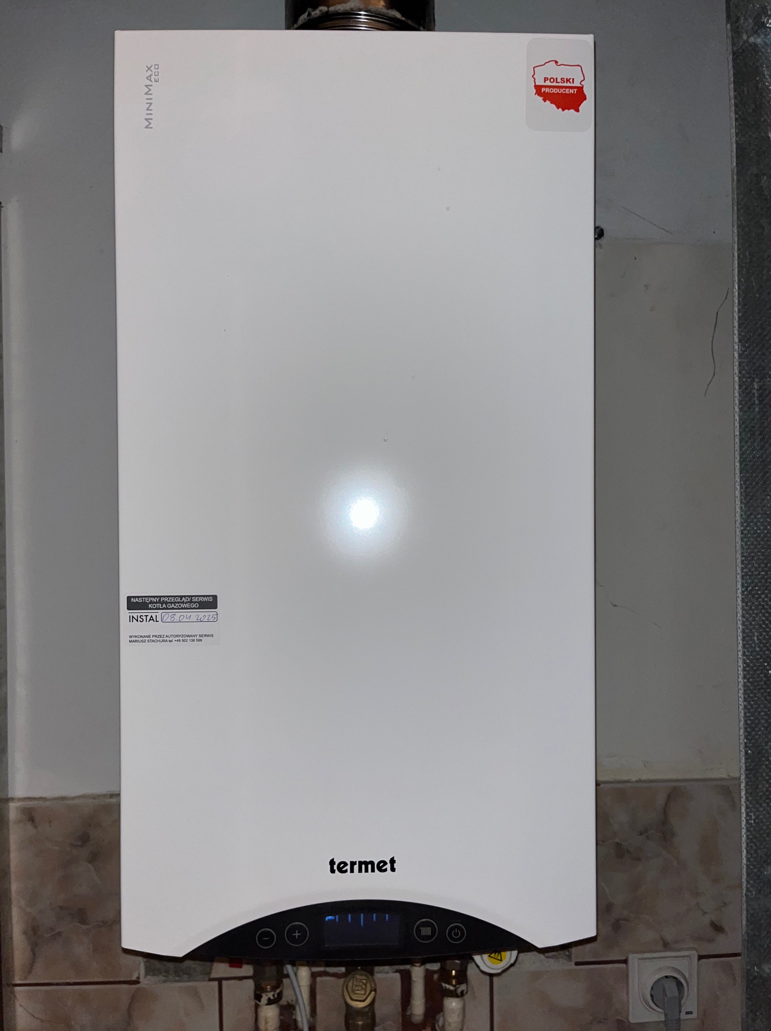 Biały piec gazowy Termet Minimax Eco zamontowany na ścianie z brązowymi płytkami, widoczna naklejka 'Polski Producent' i informacja o dacie kolejnego przeglądu.