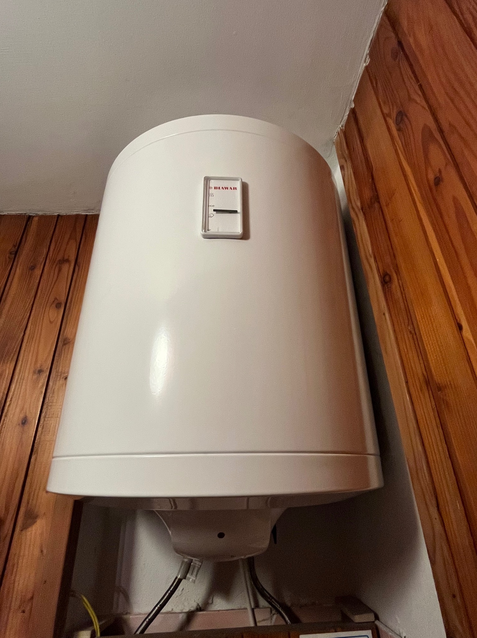 Biały, cylindryczny bojler elektryczny marki Biawar, zamontowany w rogu pomieszczenia wykończonego boazerią i białą ścianą, widoczny termostat z zakresem temperatur.