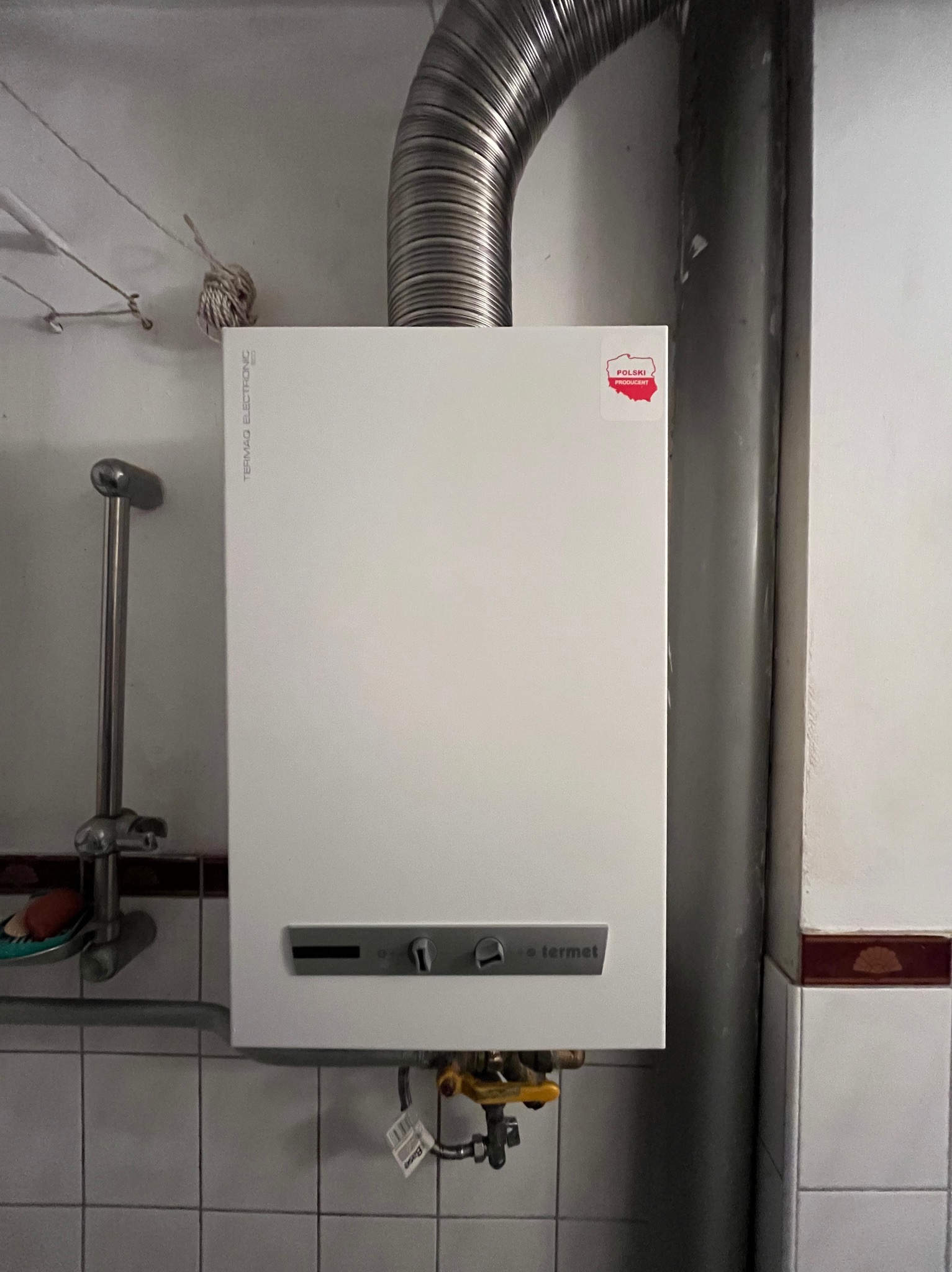 Biały piec gazowy Termet z podłączonym kominem, zamontowany na kafelkowej ścianie w łazience w Krakowie. Na piecu naklejka 'Polski Producent'. Widoczne zawory i elementy instalacji gazowej.