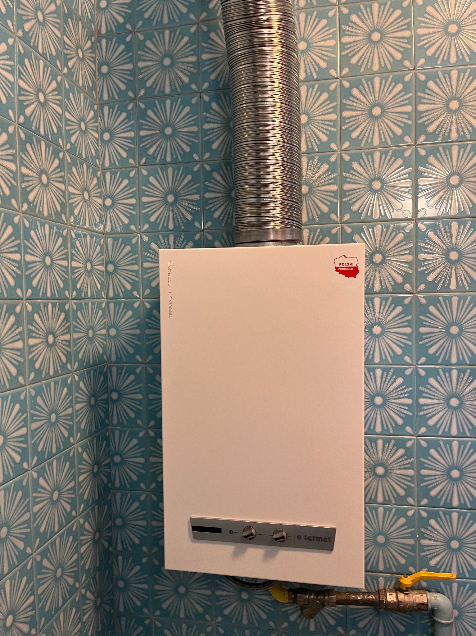 Zamontowany biały piec gazowy Termaq Electronic z kominem, na ścianie z niebieskich płytek w kwiaty, widoczny zawór gazowy z żółtą rączką.