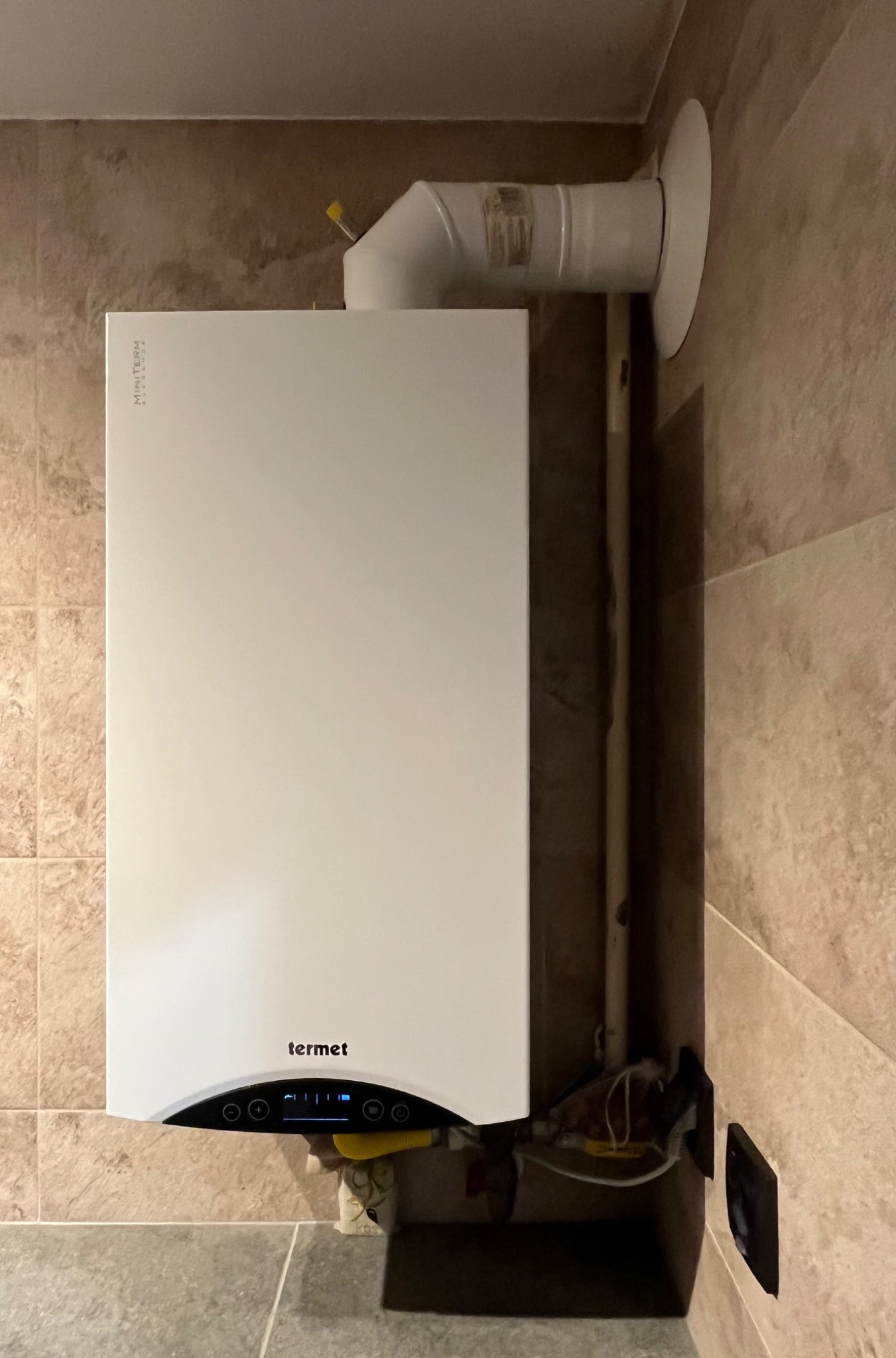 Biały piec gazowy marki Termet zamontowany na ścianie wyłożonej płytkami imitującymi kamień, widoczne rury odprowadzające spaliny i panel sterowania z wyświetlaczem.