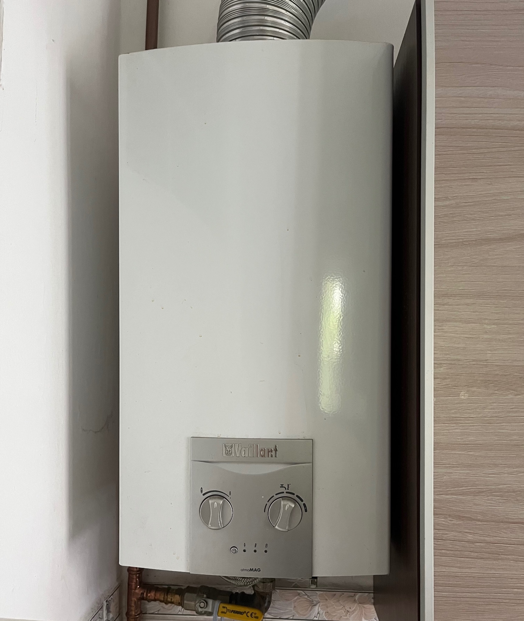 Biały piec gazowy Vaillant zamontowany na ścianie, widoczne rury miedziane i zawór gazowy, obok szafka z drewnopodobnym wykończeniem.