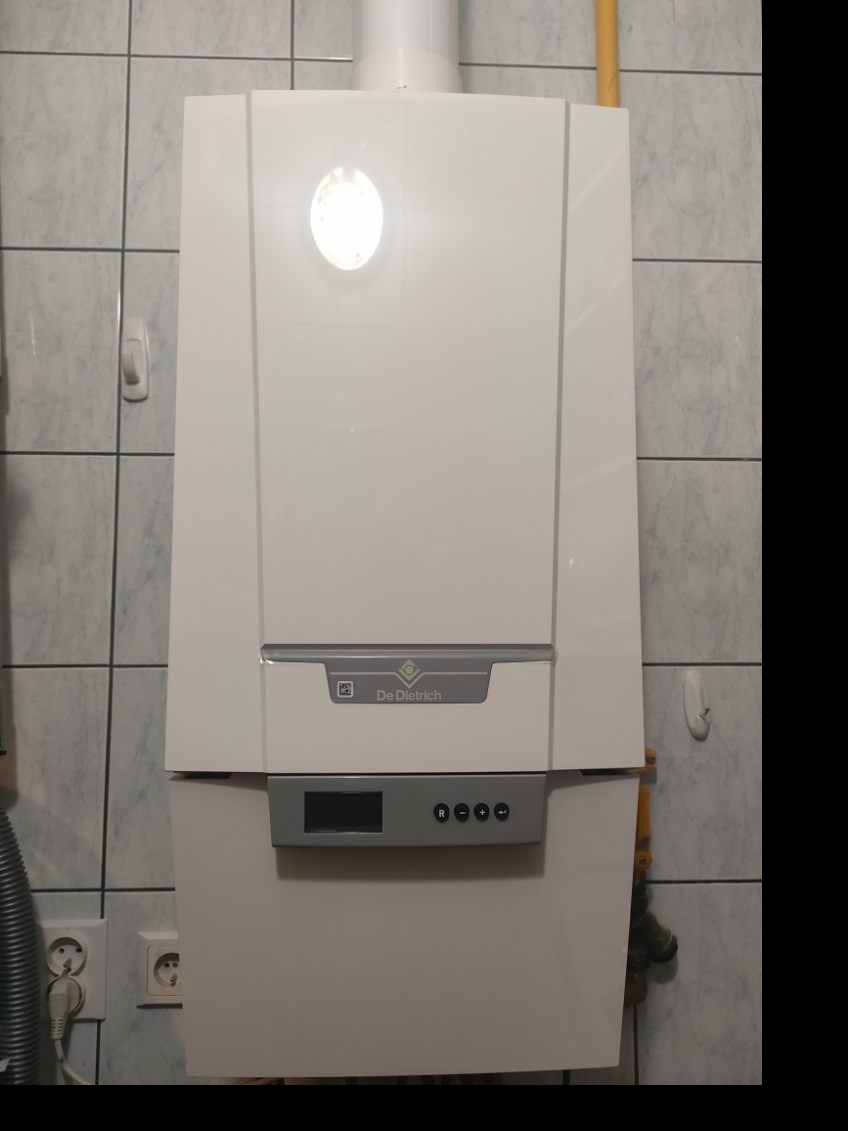 Biały piec gazowy De Dietrich zamontowany na ścianie z jasnych, kwadratowych płytek. Widoczny przewód gazowy i gniazdko elektryczne obok.