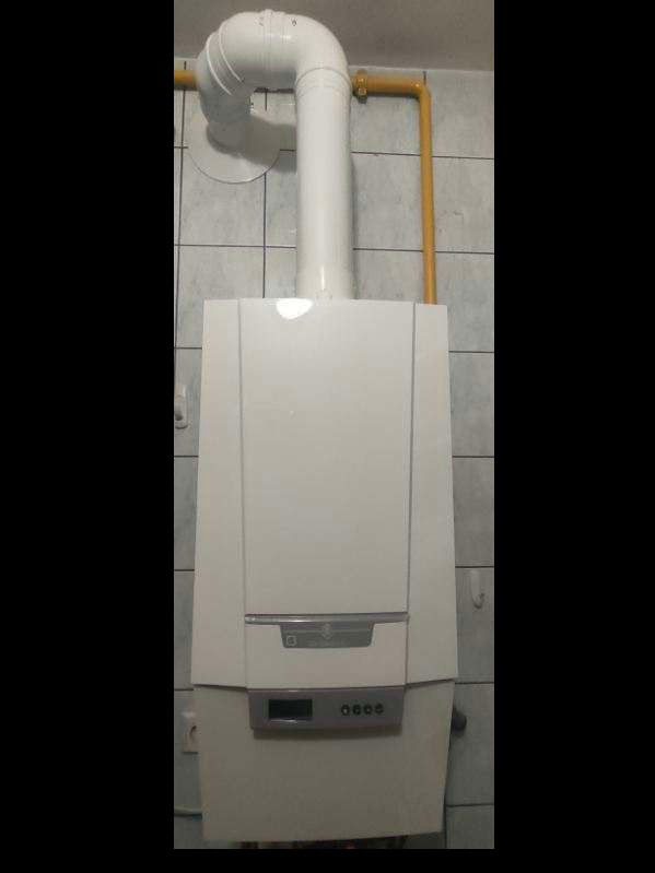 Zamontowany, biały piec gazowy z białym kominem i żółtą rurą gazową na tle szarych, kwadratowych płytek.