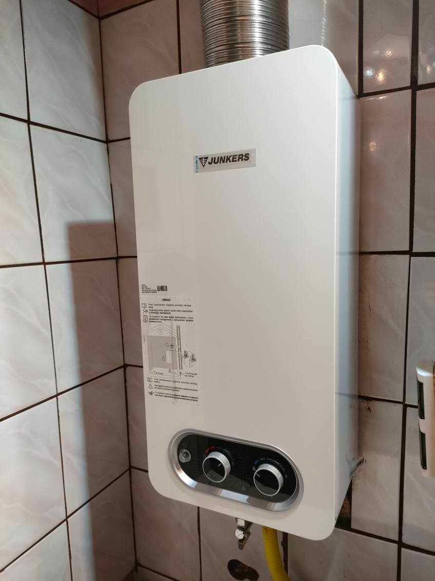 Biały piec gazowy Junkers zamontowany na ścianie wyłożonej kafelkami, z widoczną żółtą rurą gazową i elastycznym przewodem spalinowym.