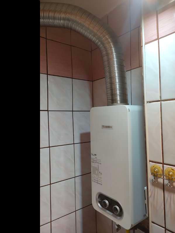 Biały piec gazowy z podłączonym, elastycznym przewodem spalinowym w łazience wyłożonej biało-różowymi płytkami.