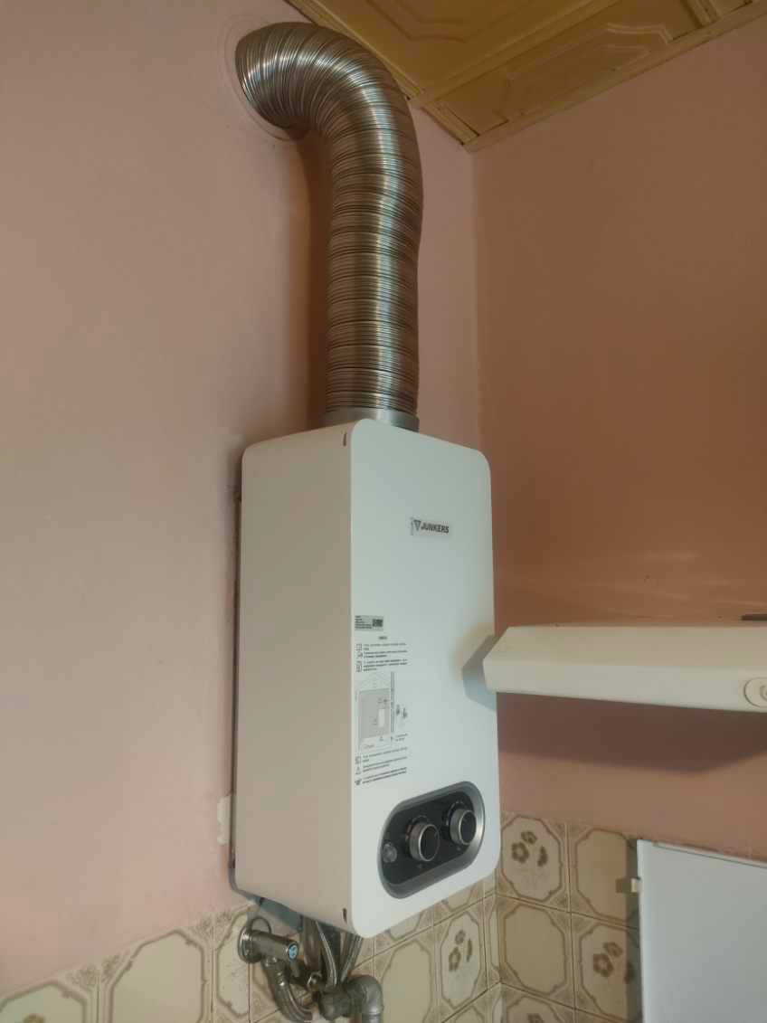 Biały piec gazowy Junkers z podłączonym, karbowanym kominem odprowadzającym spaliny, zamontowany na różowej ścianie w Krakowie, nad płytkami ceramicznymi.