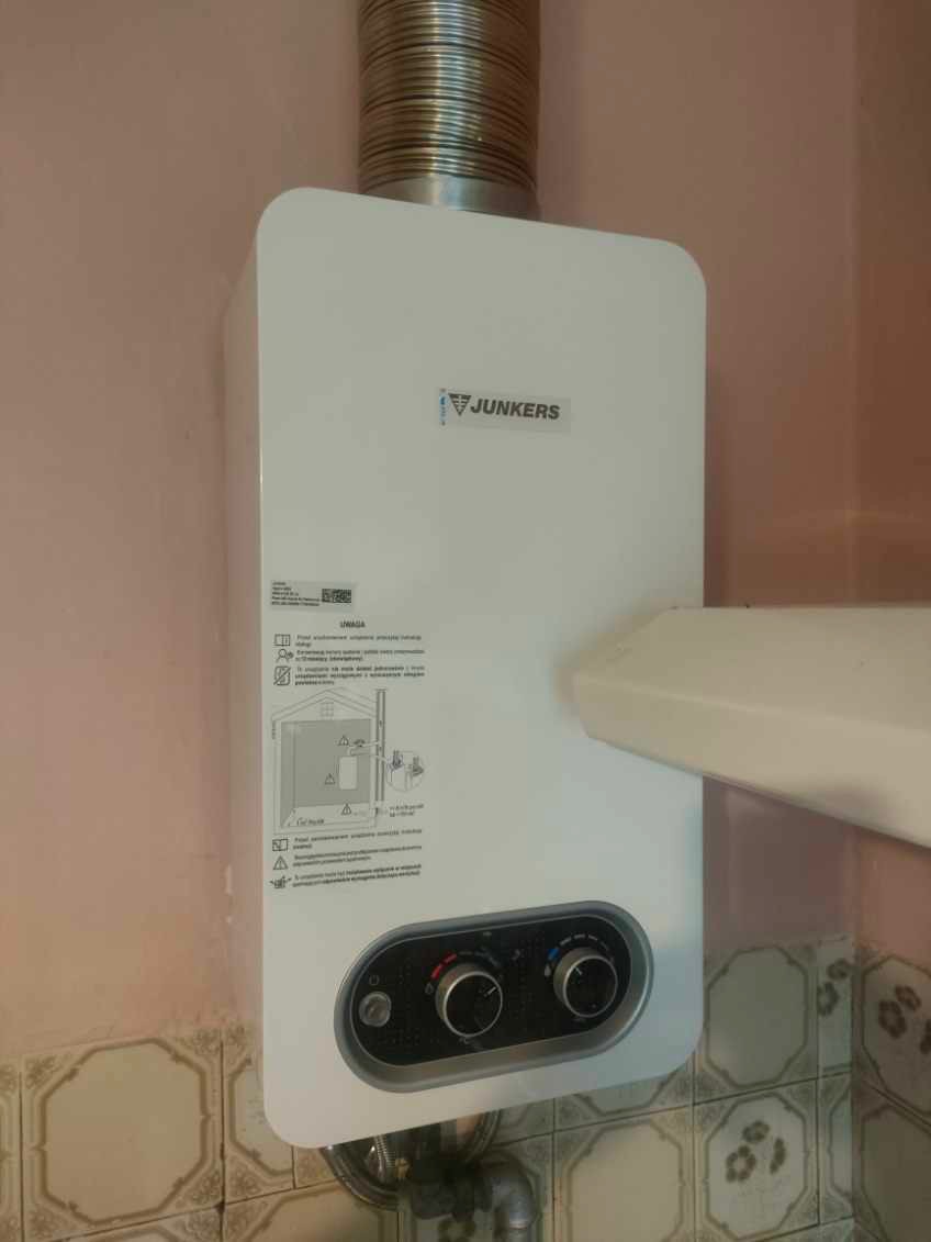 Biały piec gazowy Junkers z podłączonym kominem i widocznymi pokrętłami regulacji, zamontowany na ścianie wyłożonej płytkami w odcieniach beżu i brązu.
