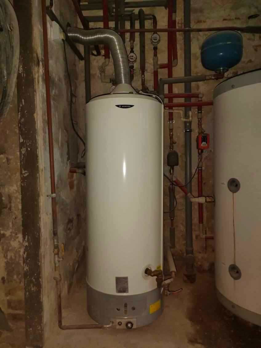 Biały piec gazowy Ariston z podłączonymi rurami w piwnicy o ścianach z cegły, widoczny także drugi zbiornik i instalacja hydrauliczna.