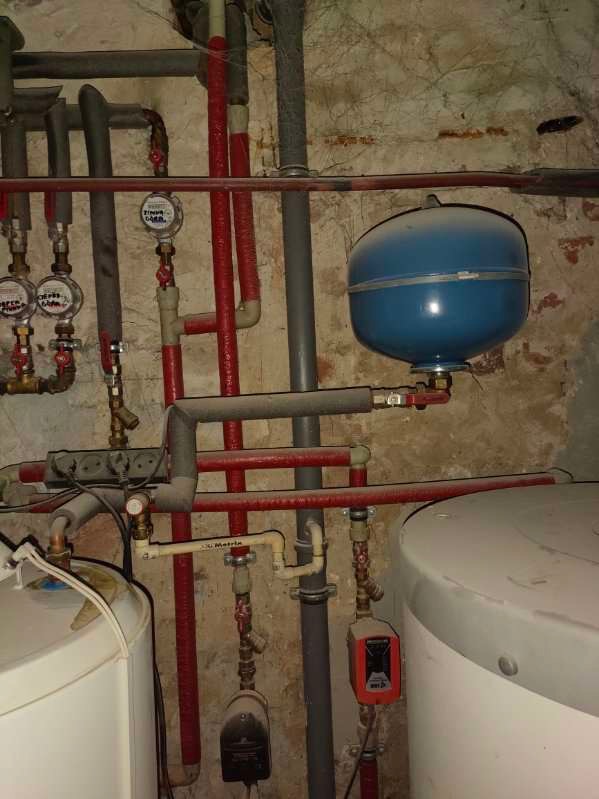 Węzeł instalacji centralnego ogrzewania z widocznym zbiornikiem wyrównawczym, rurami w izolacji i elementami armatury na tle surowej, ceglanej ściany w piwnicy.