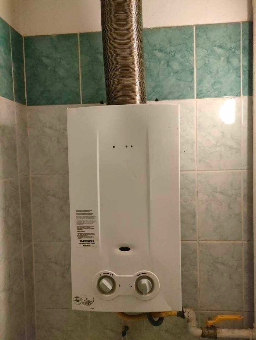 Biały piec gazowy Junkers zamontowany na ścianie z zielonymi płytkami, z podłączonym metalowym kominem spalinowym.