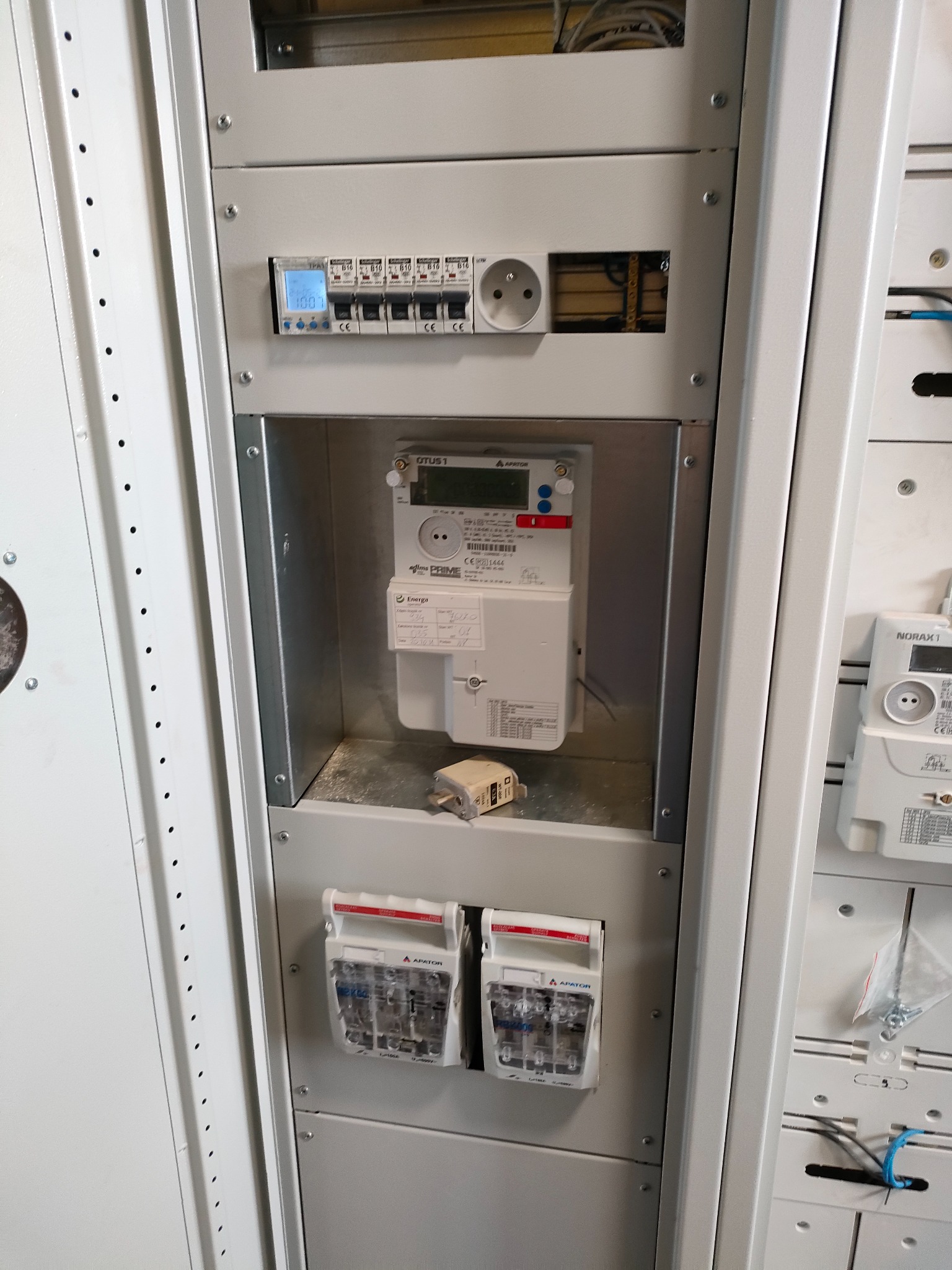 Wnętrze rozdzielnicy elektrycznej z zamontowanym licznikiem energii elektrycznej i zabezpieczeniami nadprądowymi, widoczne przewody i gniazdo zasilające.