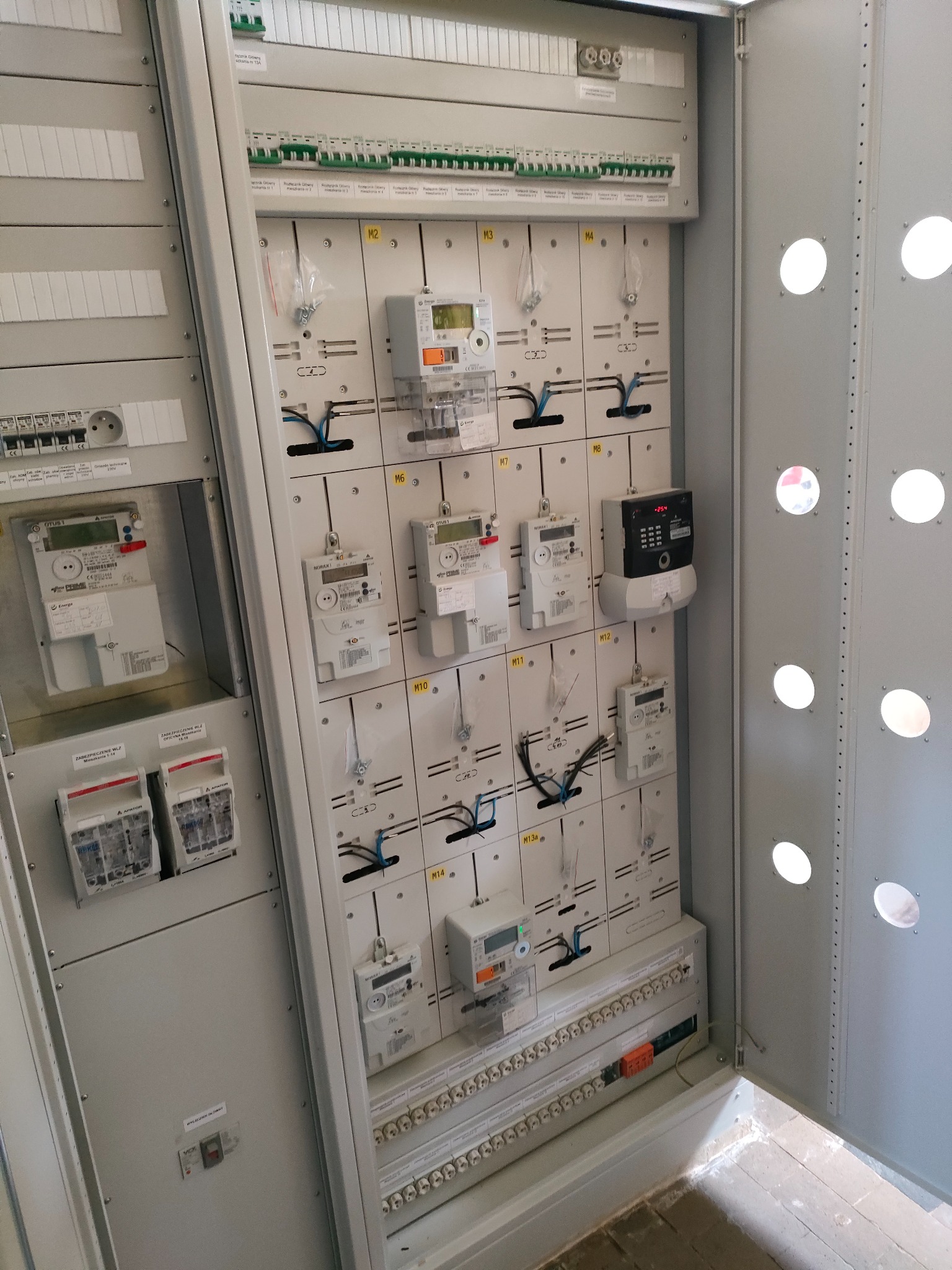 Otwarta szafa rozdzielcza z zamontowanymi licznikami energii elektrycznej, bezpiecznikami i listwami zaciskowymi, widoczne okablowanie i oznaczenia poszczególnych modułów.