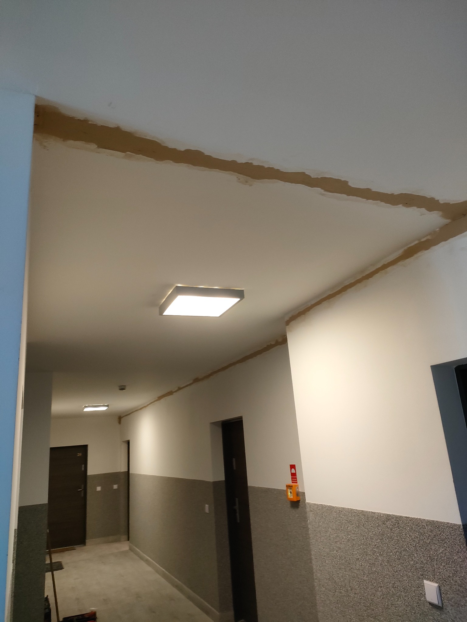 Korytarz w budynku wielorodzinnym z widocznymi śladami po prowadzeniu instalacji pod sufitem, z zamontowanym oświetleniem LED i czujnikiem dymu.