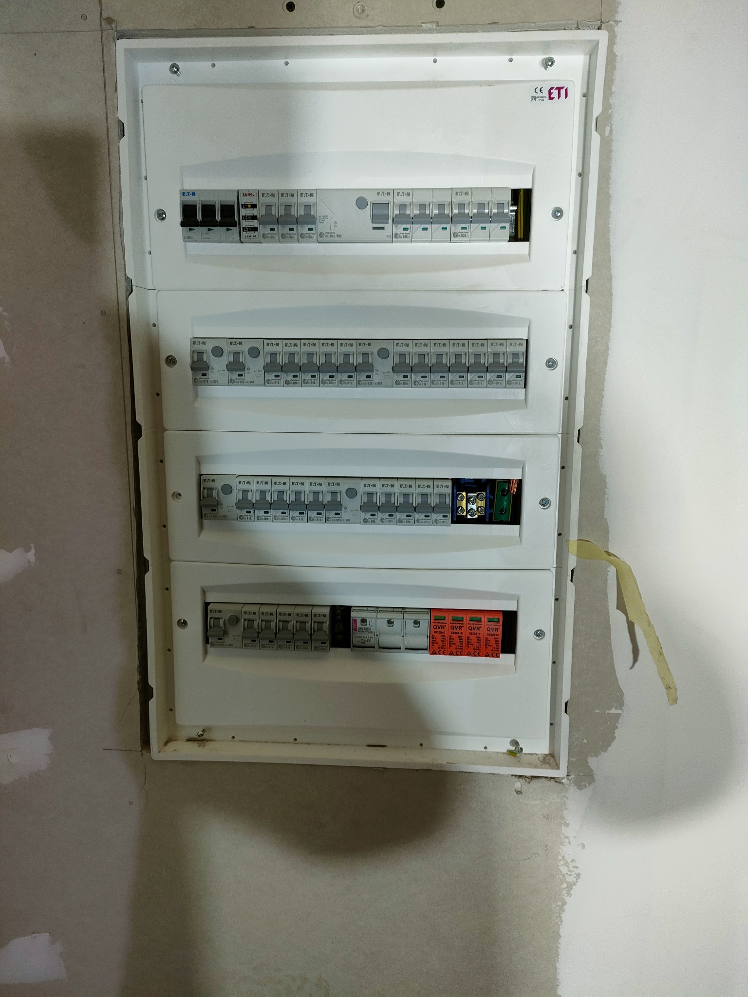 Rozdzielnica elektryczna z zabezpieczeniami różnicowoprądowymi i nadprądowymi, zamontowana na surowej ścianie z widocznymi śladami gładzi szpachlowej i taśmy malarskiej.