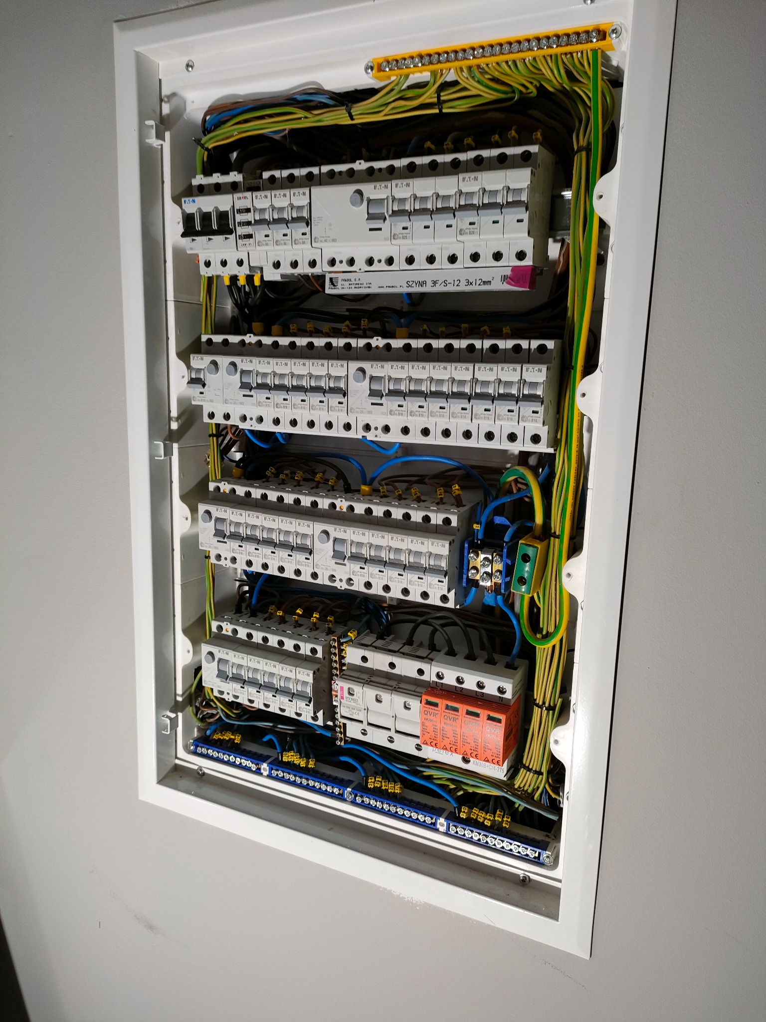 Rozdzielnia elektryczna w białej obudowie z widocznymi bezpiecznikami, przewodami w kolorach niebieskim, żółto-zielonym i brązowym, listwami zaciskowymi i zabezpieczeniami przepięciowymi marki...