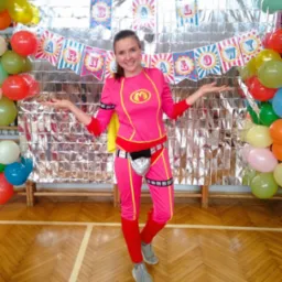 Animator w różowym kostiumie superbohatera z literą 'M' na tle srebrnej ściany z balonami i napisem 'Happy Birthday' w Orzeszu, z rozłożonymi rękami i uśmiechem.