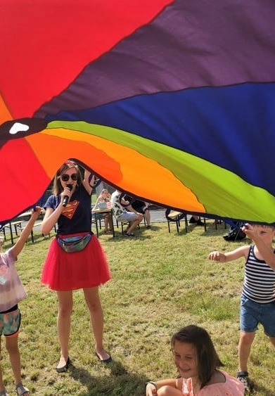 Animatorka w czerwonej spódniczce i koszulce z symbolem Supermana trzyma mikrofon pod kolorową chustą, pod którą bawią się dzieci na trawie.