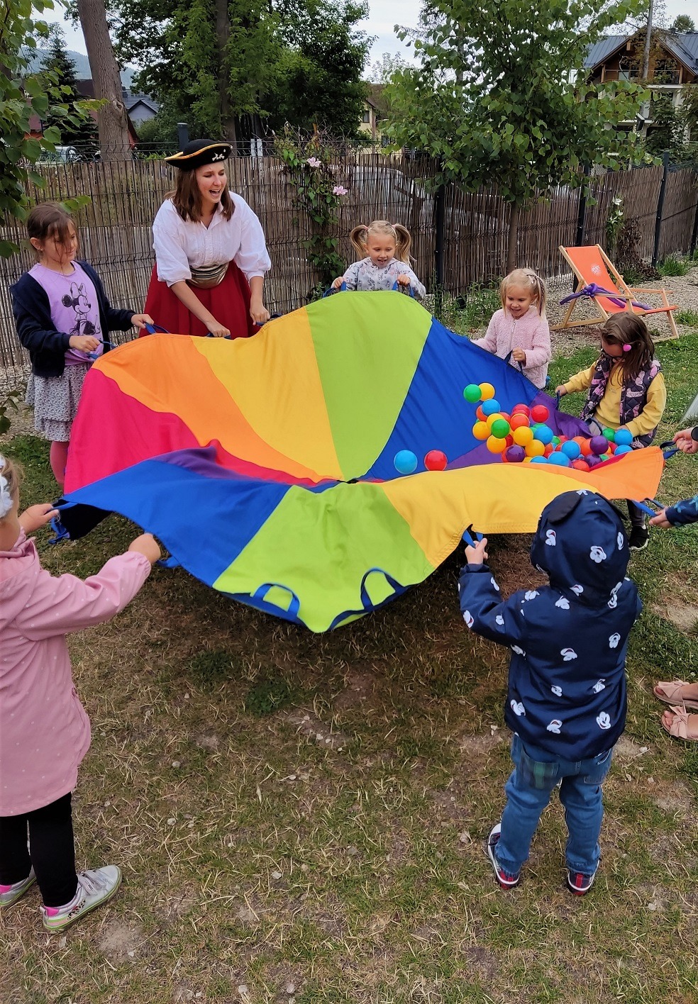 Grupa dzieci bawi się kolorową chustą animacyjną wypełnioną piłeczkami pod okiem animatorki przebranej za pirata.